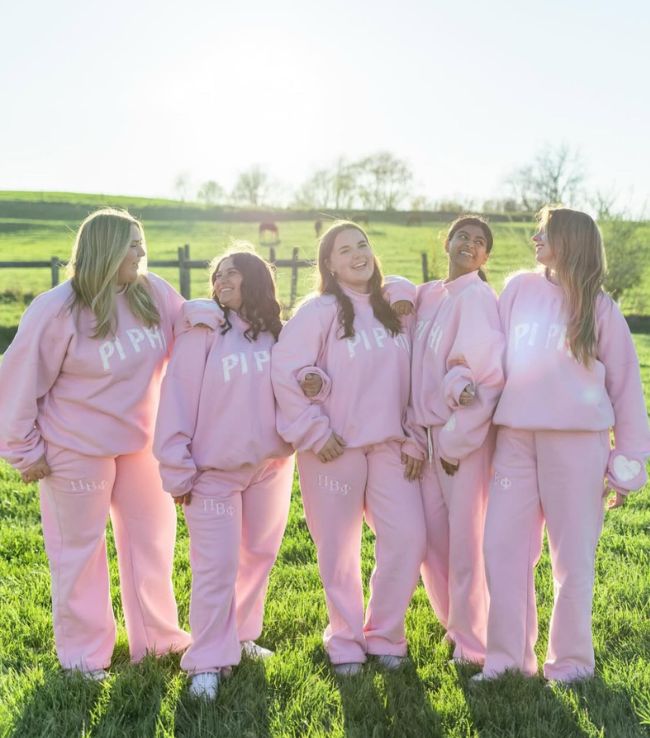 Five Pi Beta Phi members laughing outdoors in matching pink Pi Beta Phi sweatshirts and sweatpants on a grassy field