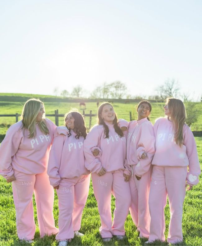 Five Pi Beta Phi members laughing together in a sunny green field wearing matching pink Pi Beta Phi sweatshirts and sweatpants