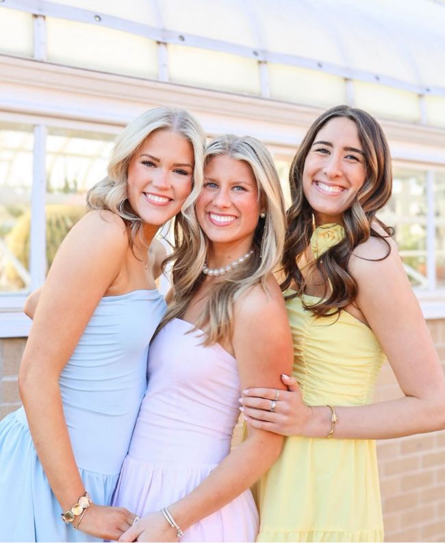 Three Pi Beta Phi members smiling together in front of a greenhouse, wearing pastel blue, pink, and yellow formal dresses