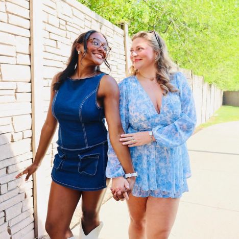 Two Pi Beta Phi members smiling and walking together outdoors on a sunny day, one wearing a denim dress with white cowboy boots and the other wearing a blue floral romper with brown cowboy boots
