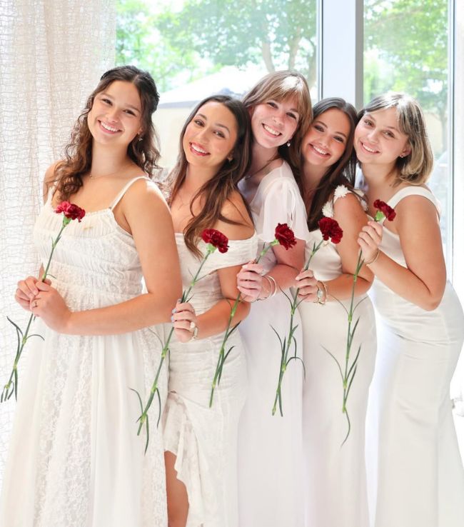 Five Pi Beta Phi members in white dresses smiling and holding red carnations indoors