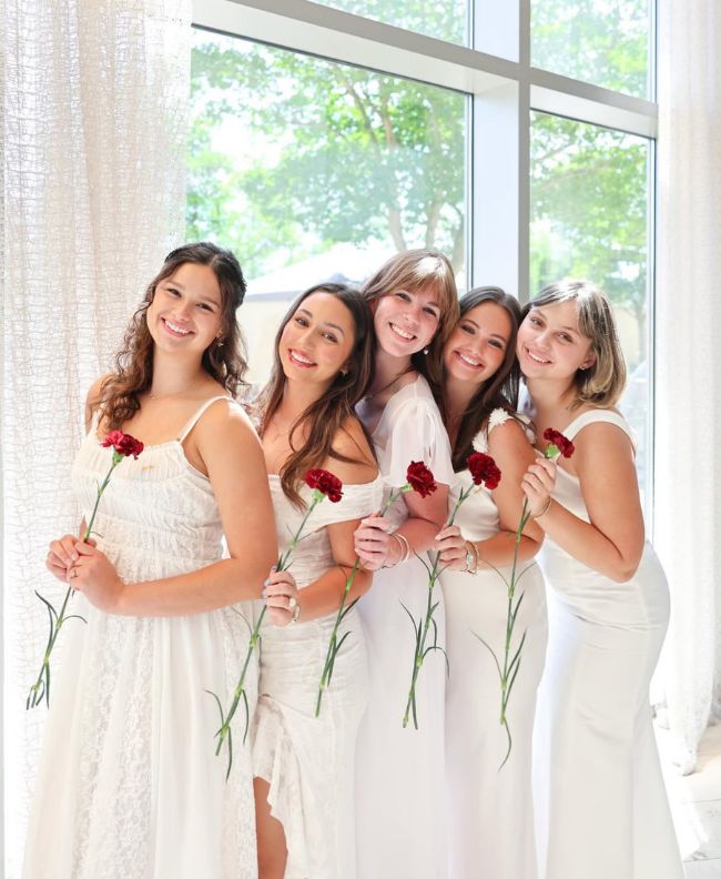 Five Pi Beta Phi members smiling indoors in white dresses, each holding a red carnation in front of a bright window