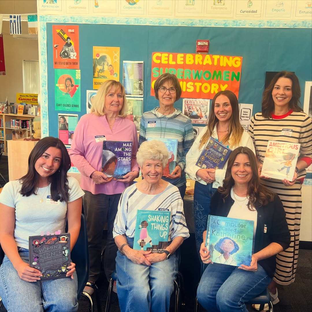 Seven alumnae members holding children's books in a classroom with a colorful bulletin board in the background