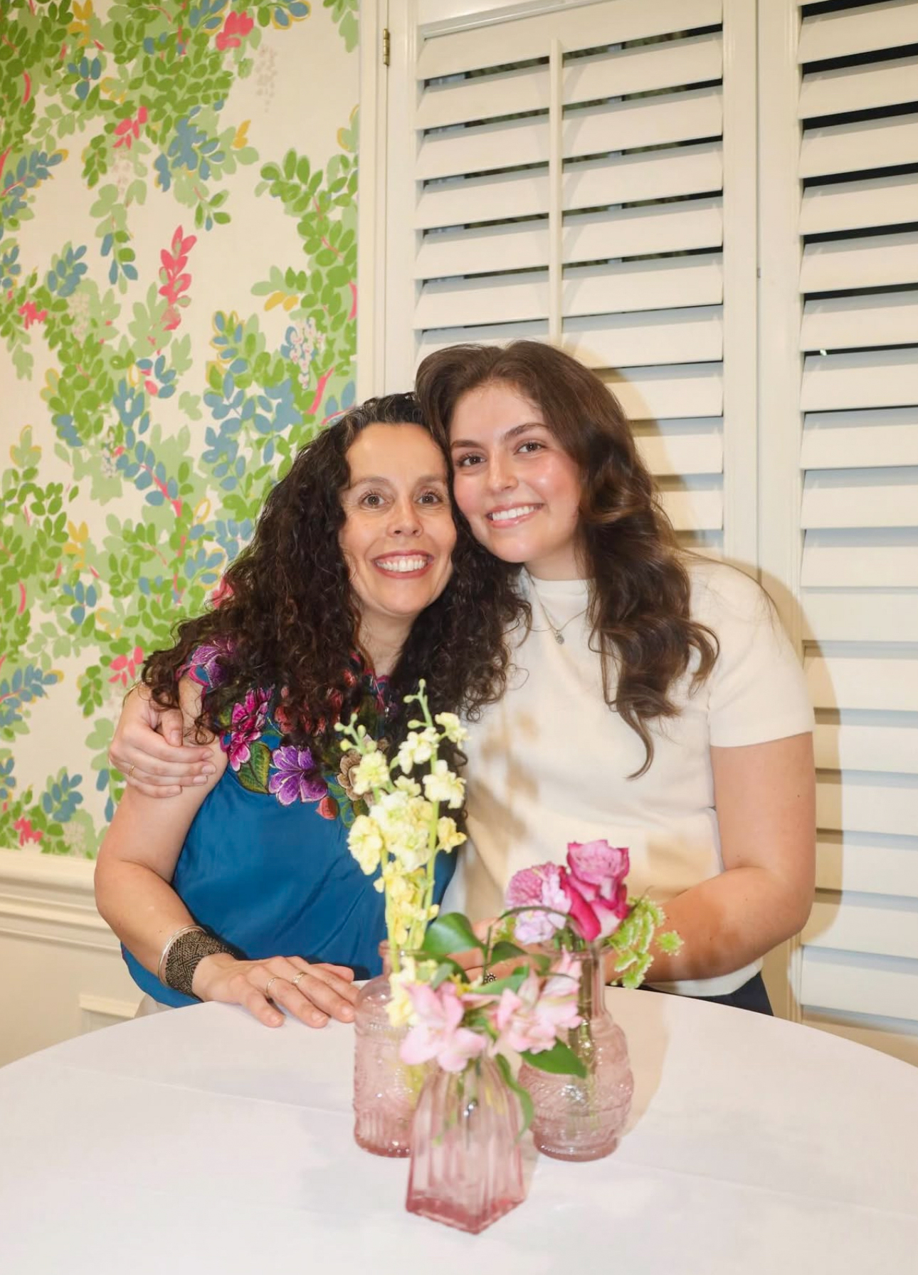 Two members sitting at a table with a white table cloth and flower centerpiece in front of a flowered wall and white windowshades