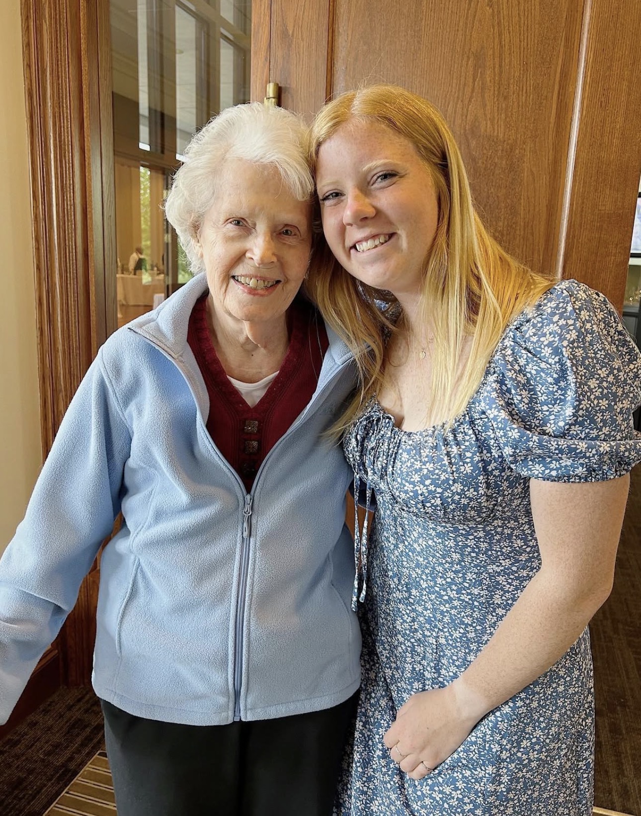 One older member in a grey jacket and one younger member in a blue flowered dress pose in front of a wood door