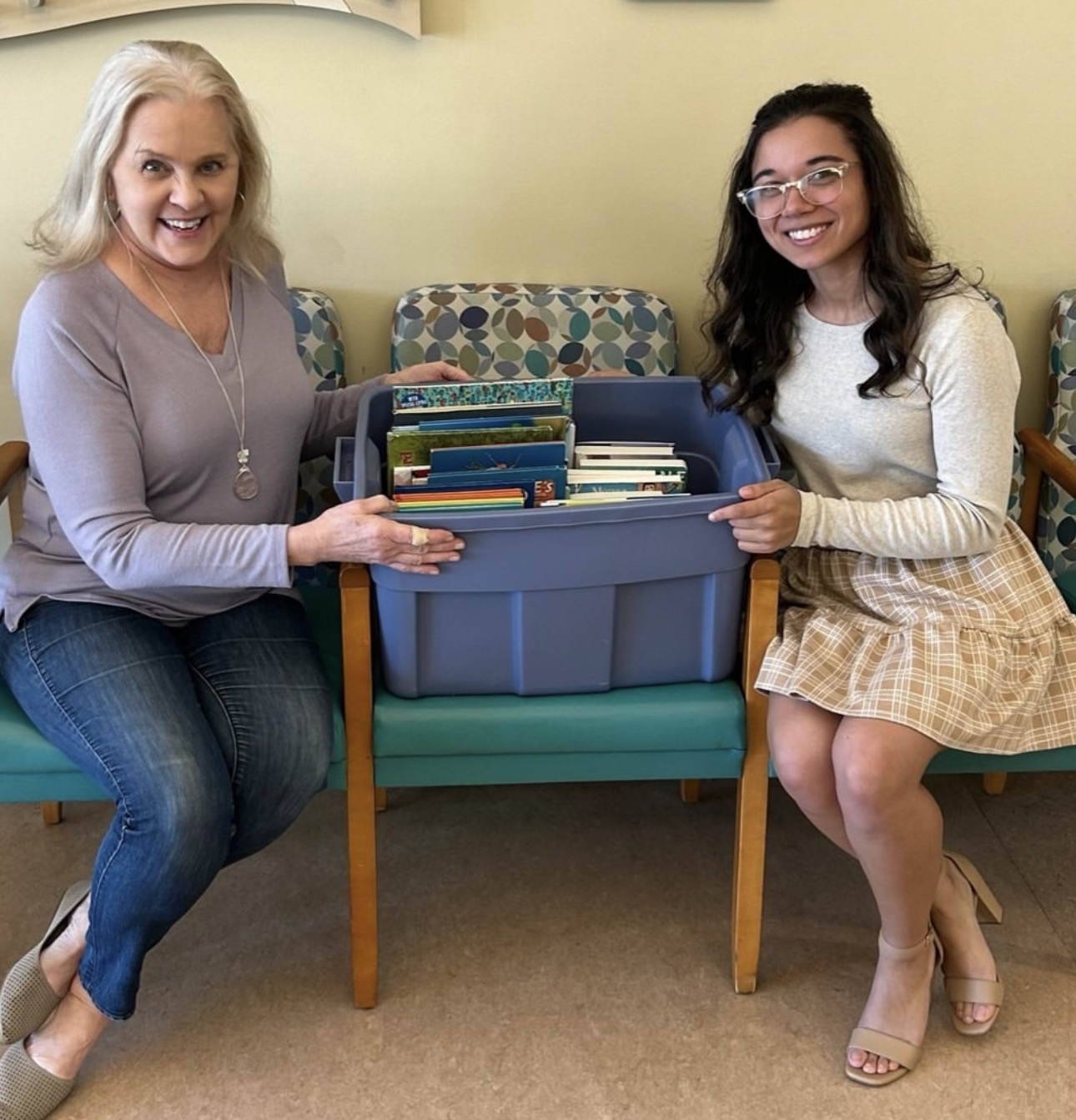 Two members sitting in chairs with a large blue bin of colorful books in between them