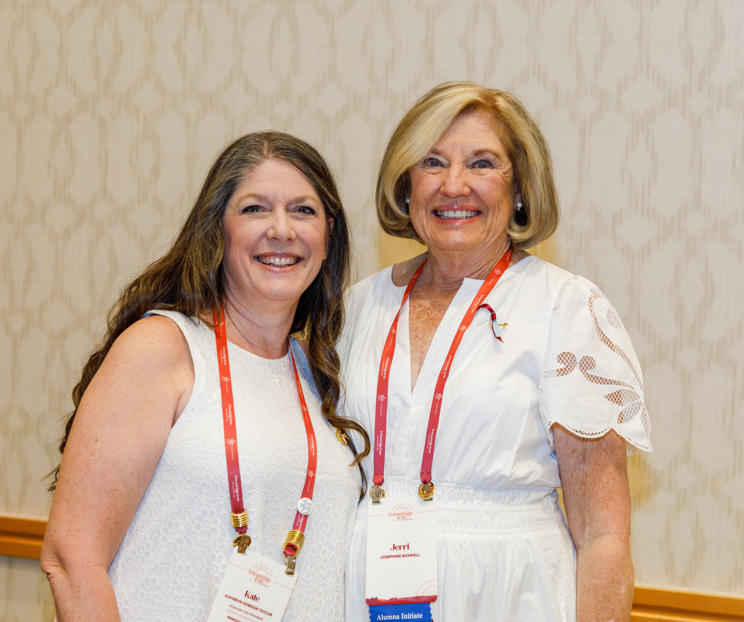 Two members wearing white dresses with red and white lanyards standing in front of a white wall
