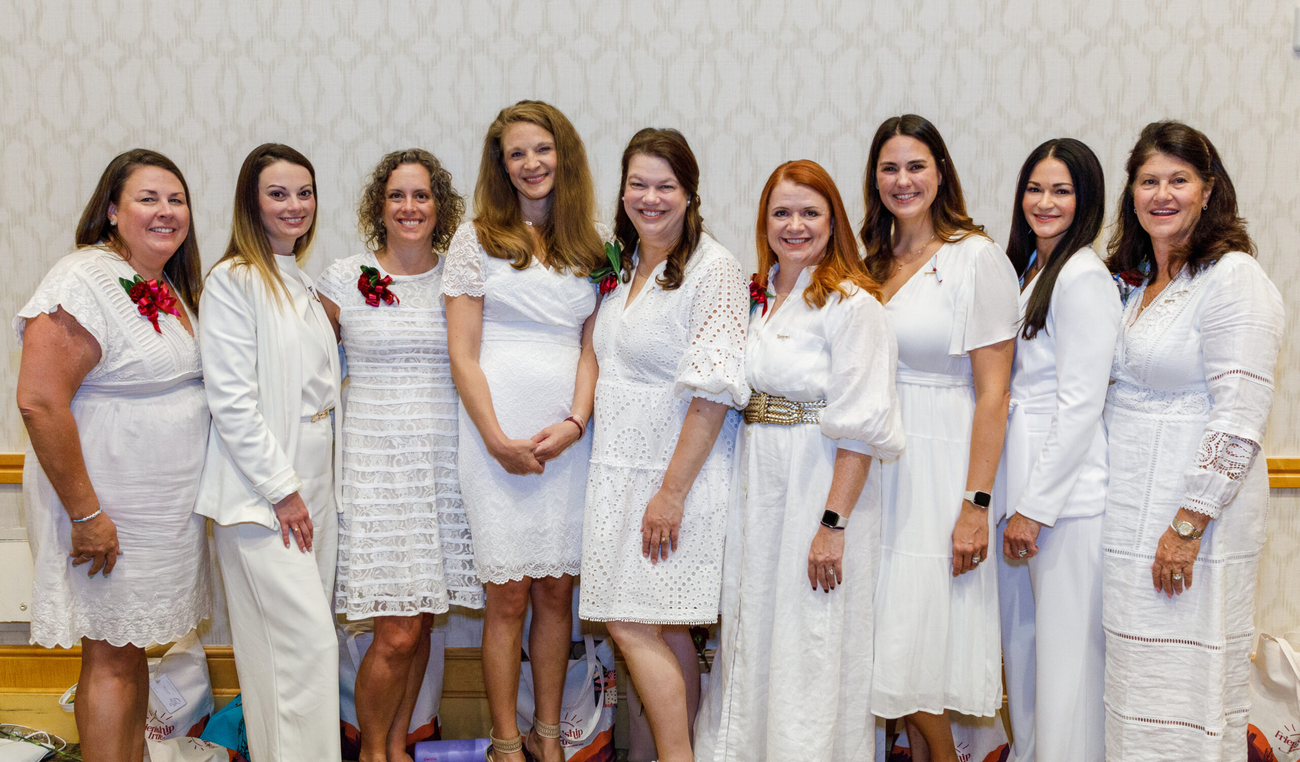 Nine alumnae wearing white dresses standing inside in front of a white wall