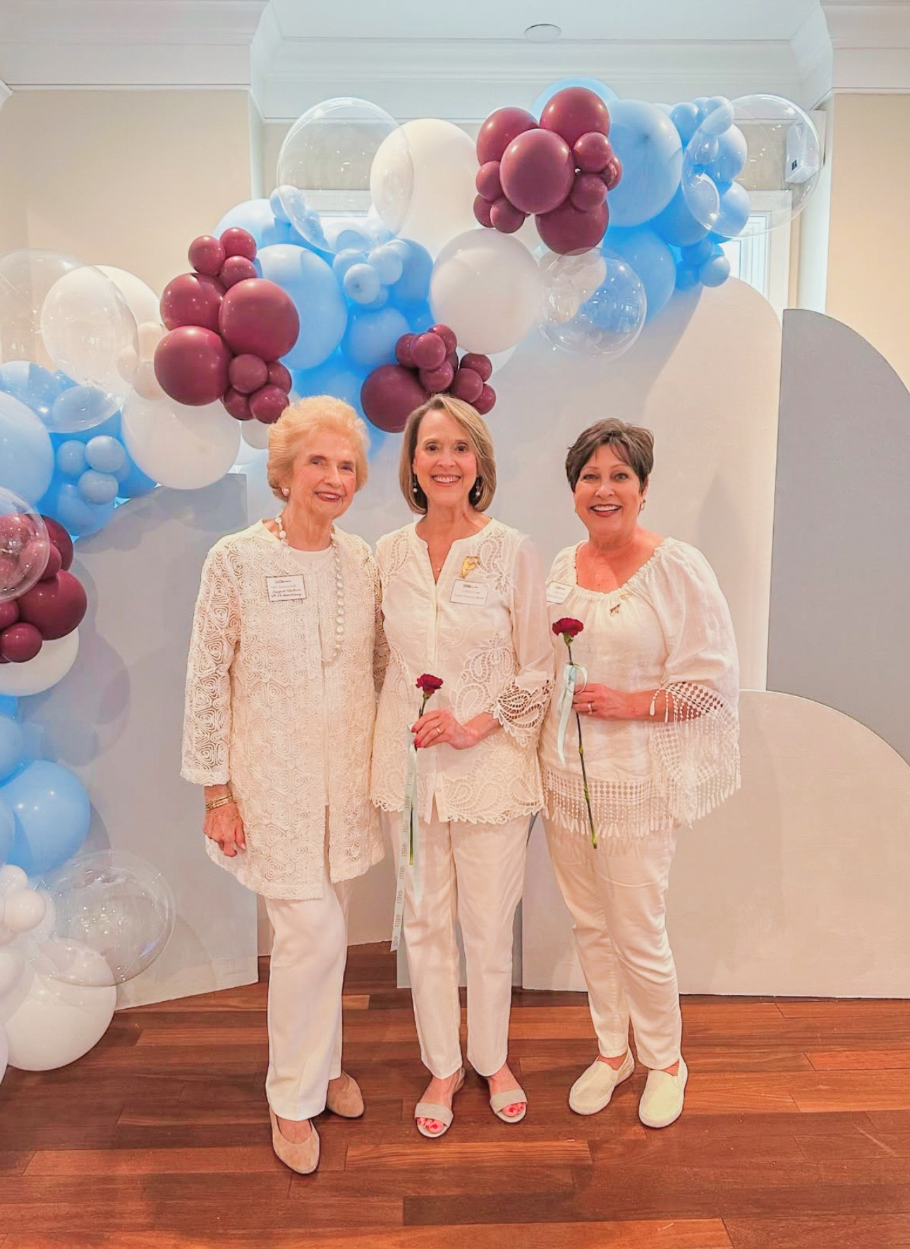 Three alumnae members wearing white shirts and white pants standing inside in front of a white background with a wine and silver blue balloon arch