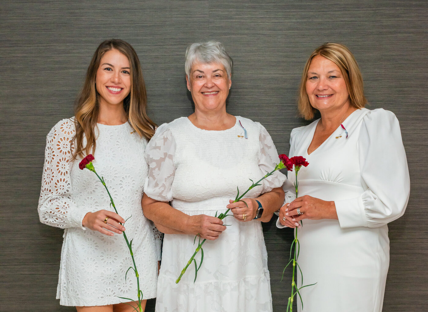 Three alumnae members wearing white dresses holding red carnations with green stems in front of a grey background