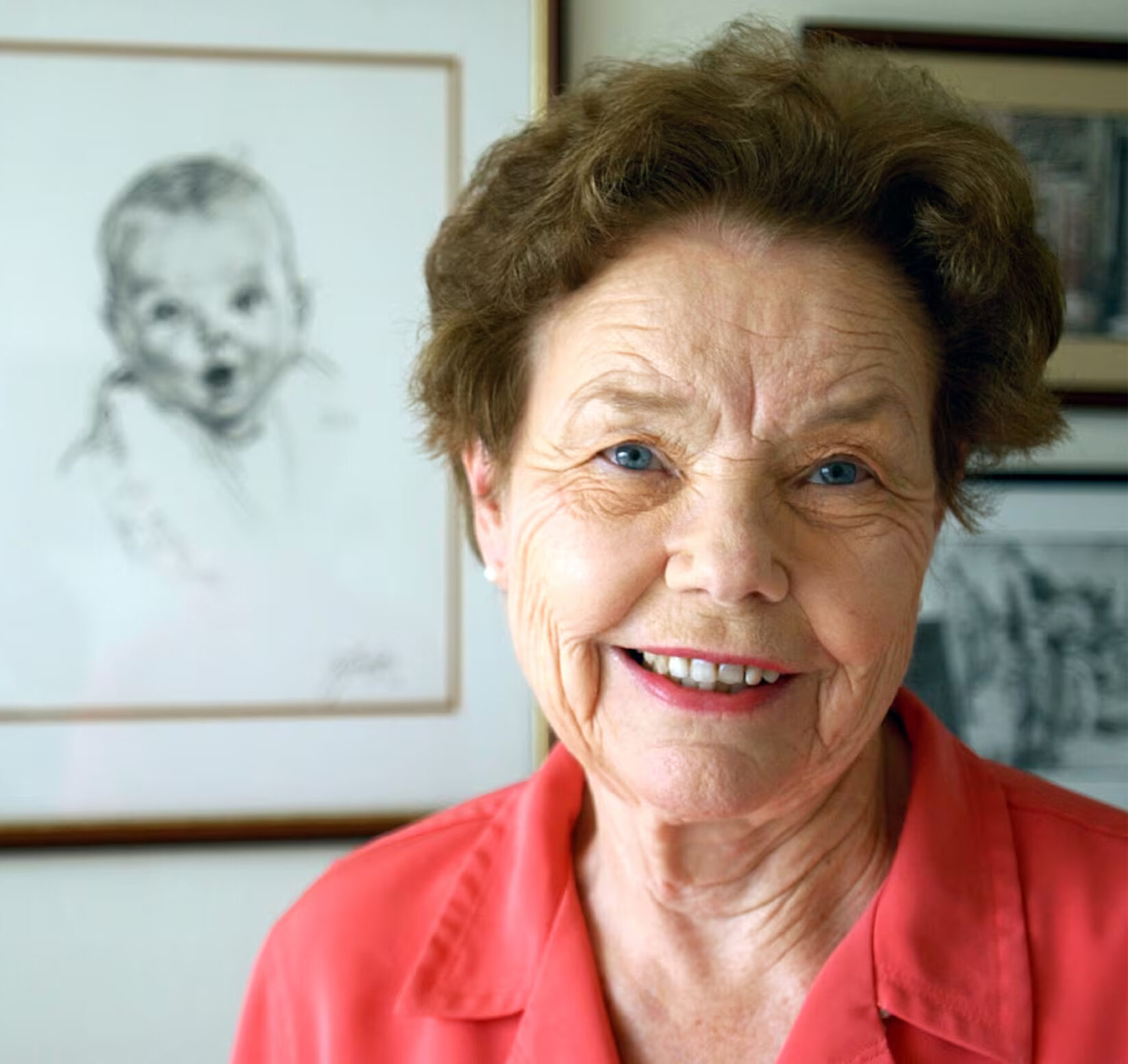 Professional headshot of Ann Cook wearing a red colored shirt posing in front of a wall with a framed photo of a baby