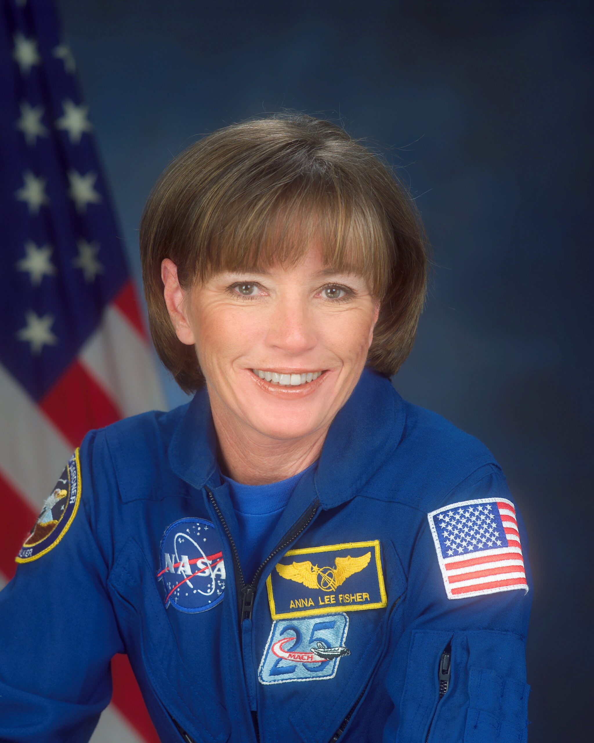 Professional headshot of Anna Lee Fisher wearing a blue NASA uniform with a yellow winged patch and American flag patch sitting in front of an American flag