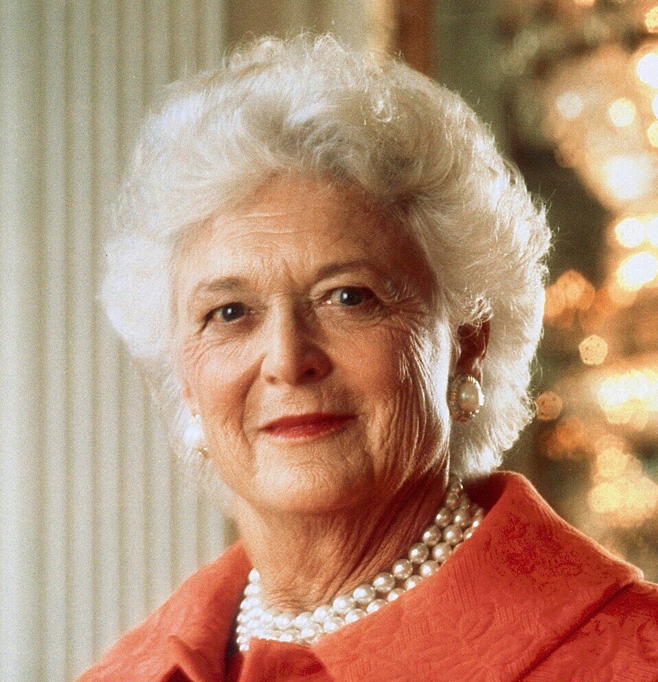 Professional headshot of Barbara Bush wearing a red blazer and pearl necklace in front of a column and lighted background