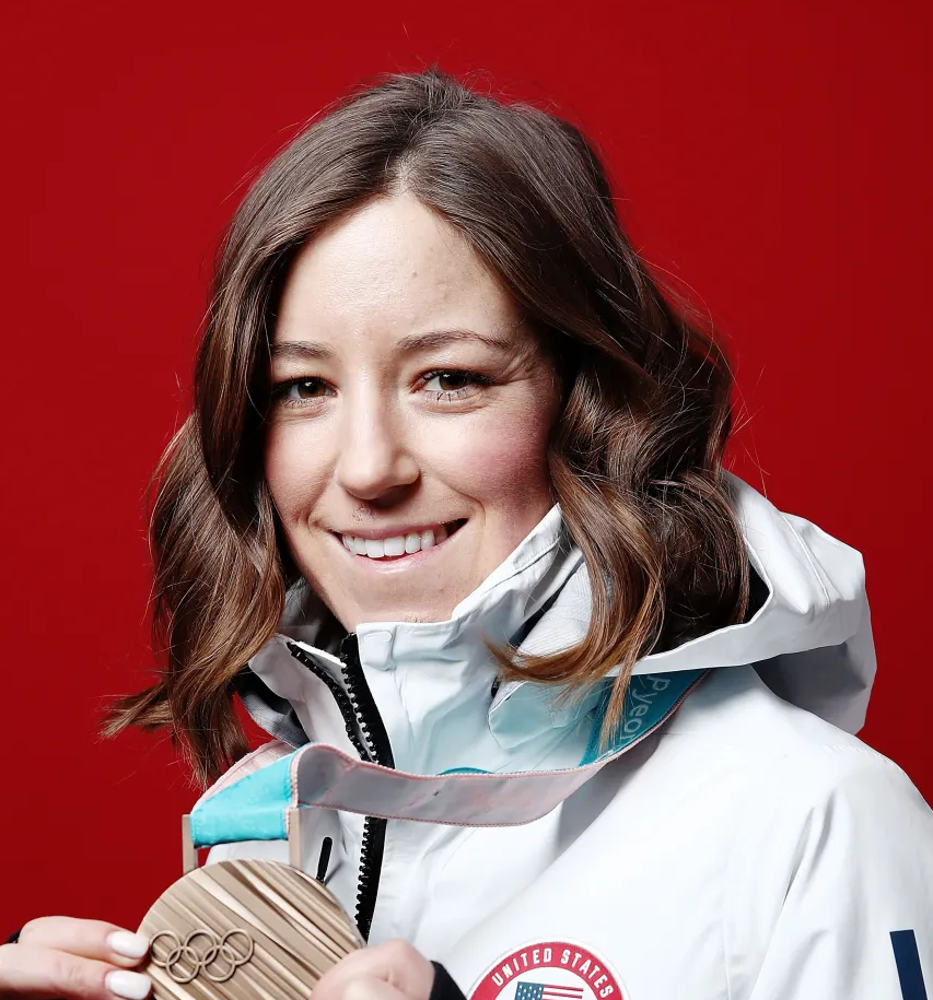 Headshot of Brita Sigourney wearing a white winter jacket holding an Olympic medal in front of a red background