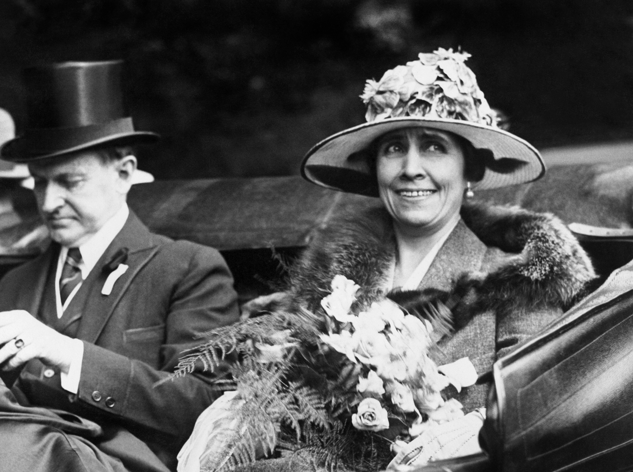 Historical black and white photo of Grace Coolidge wearing a large flowered hat and the President driving in a convertible car