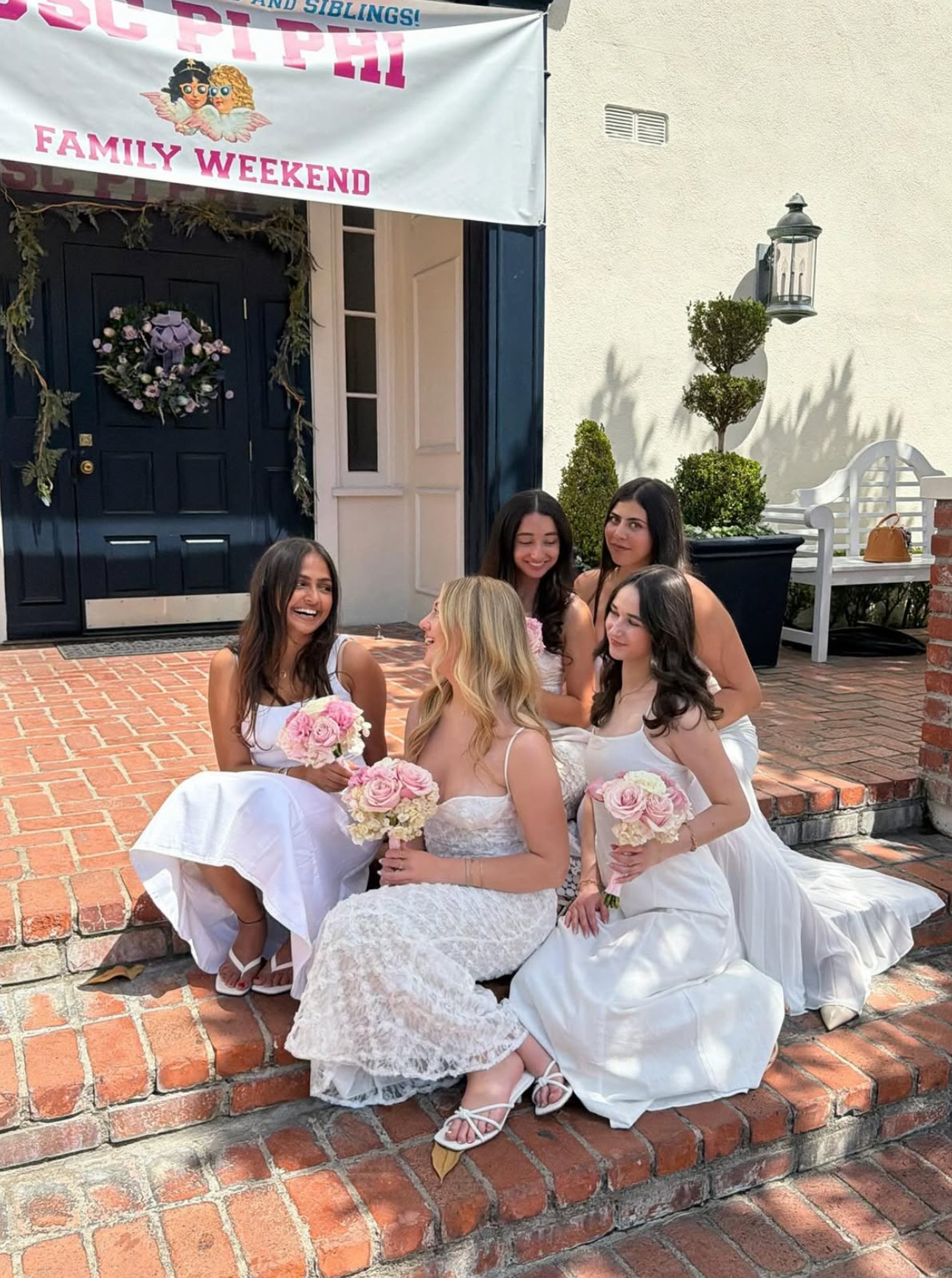 Five individuals wearing white dresses sitting on stairs outdoors