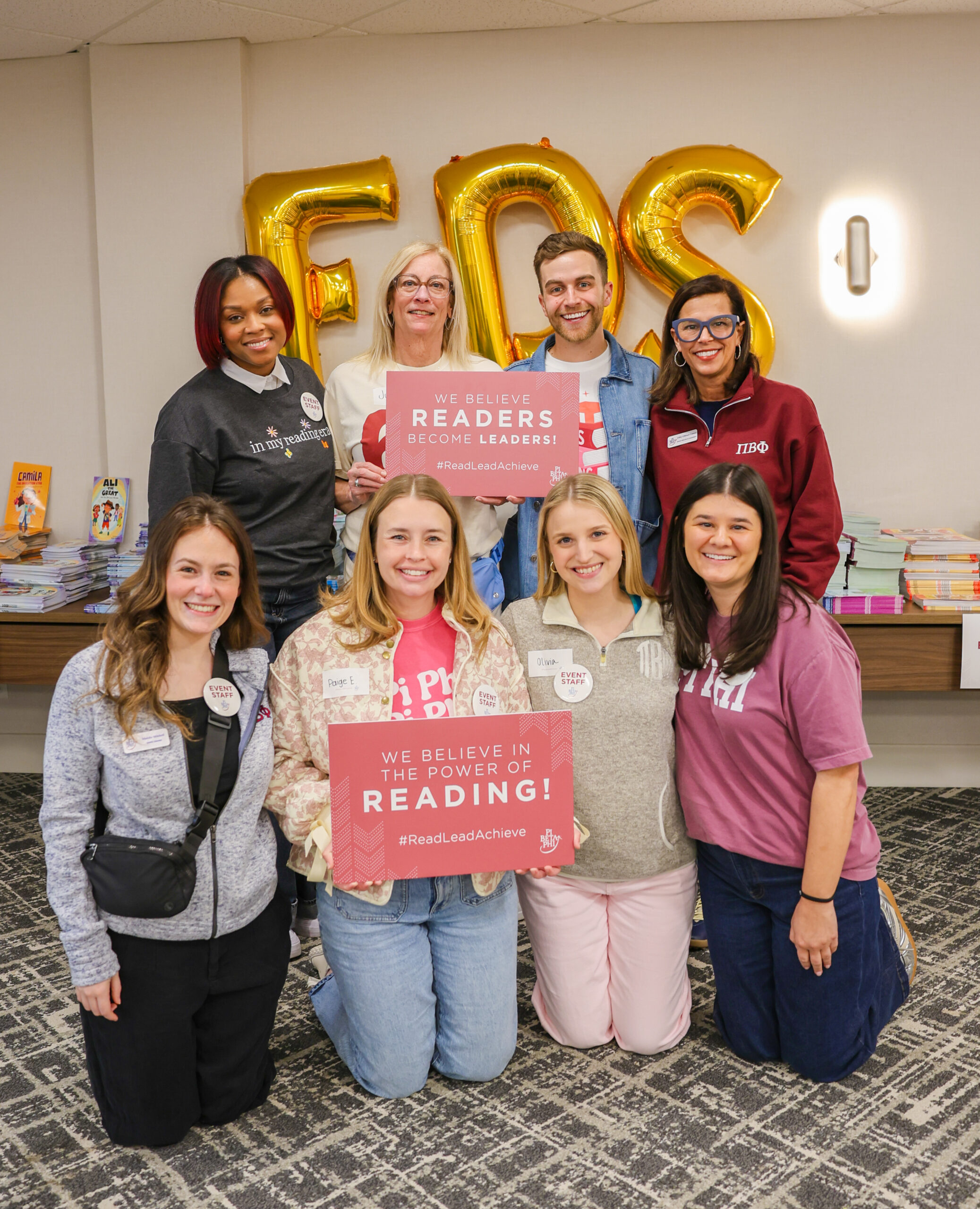 Eight Pi Beta Phi staff members posing inside holding up red signs in front of FDS gold letter balloons