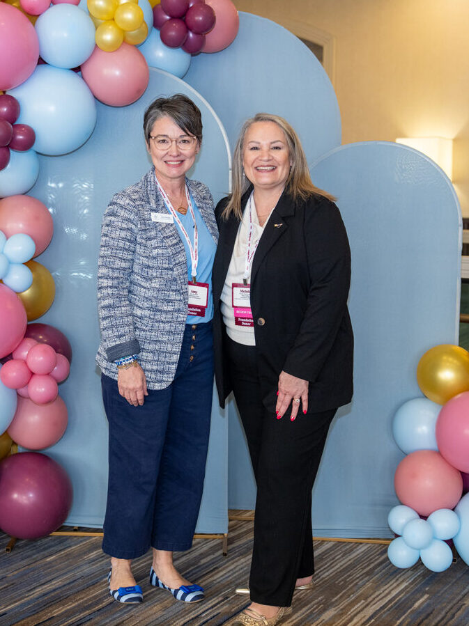 Two Pi Beta Phi staff members standing in front of a blue backdrop with wine and silver blue balloons