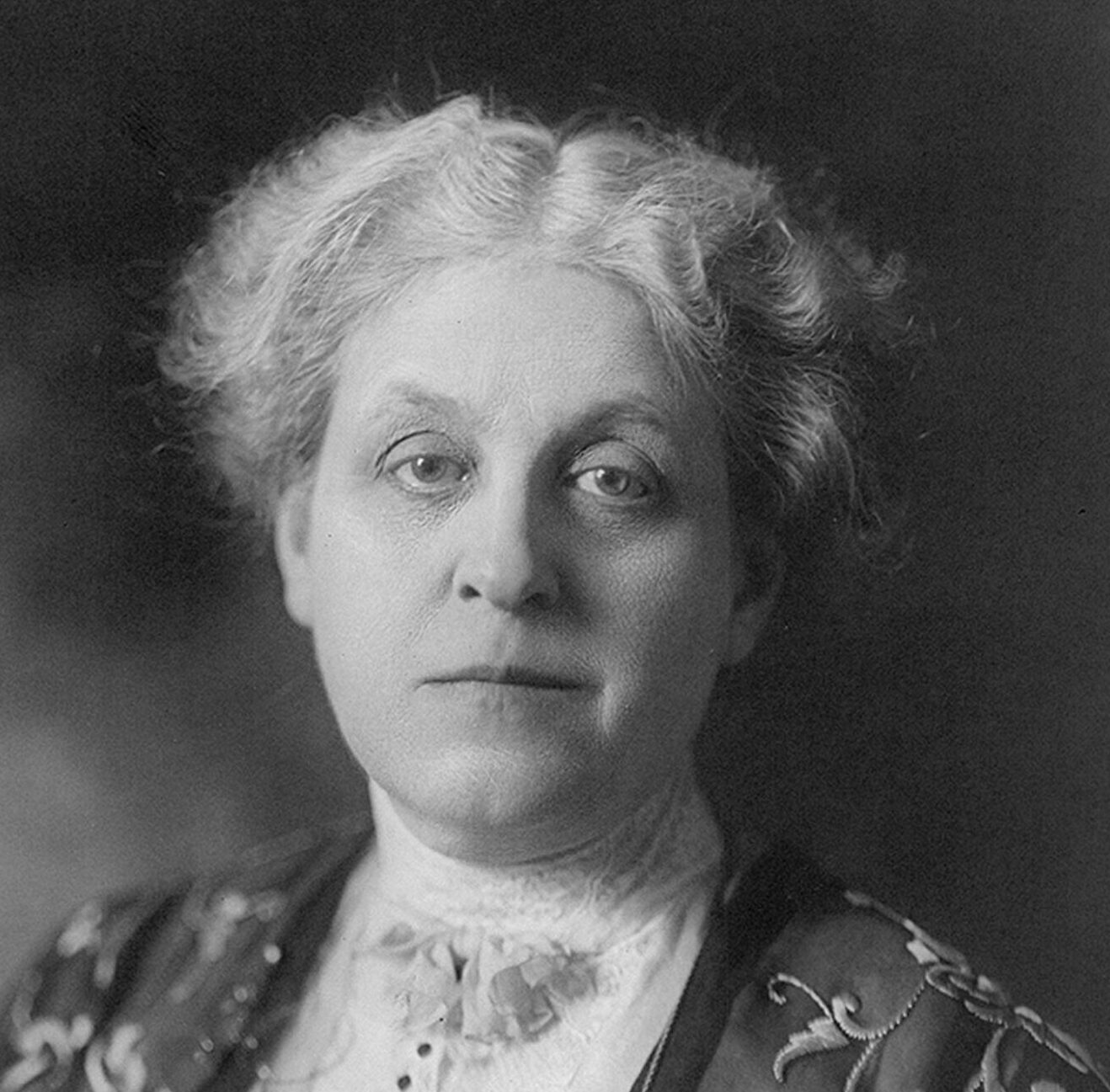 Historical black and white headshot of Carrie Chapman Catt wearing a black and white ruffled blouse in front of a black background