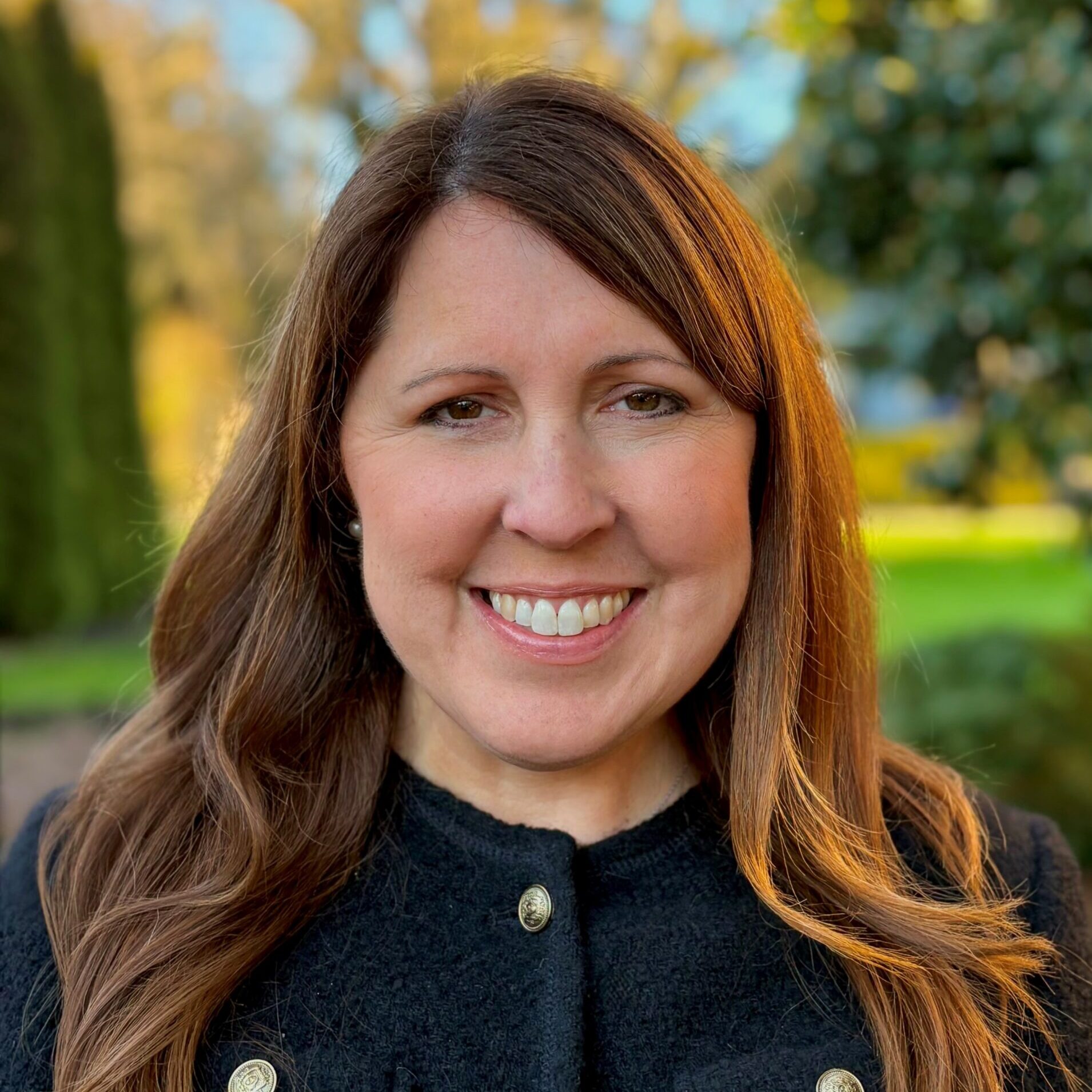 Professional headshot of Barbra Littrell wearing a black blazer with gold buttons standing in front of autumn trees