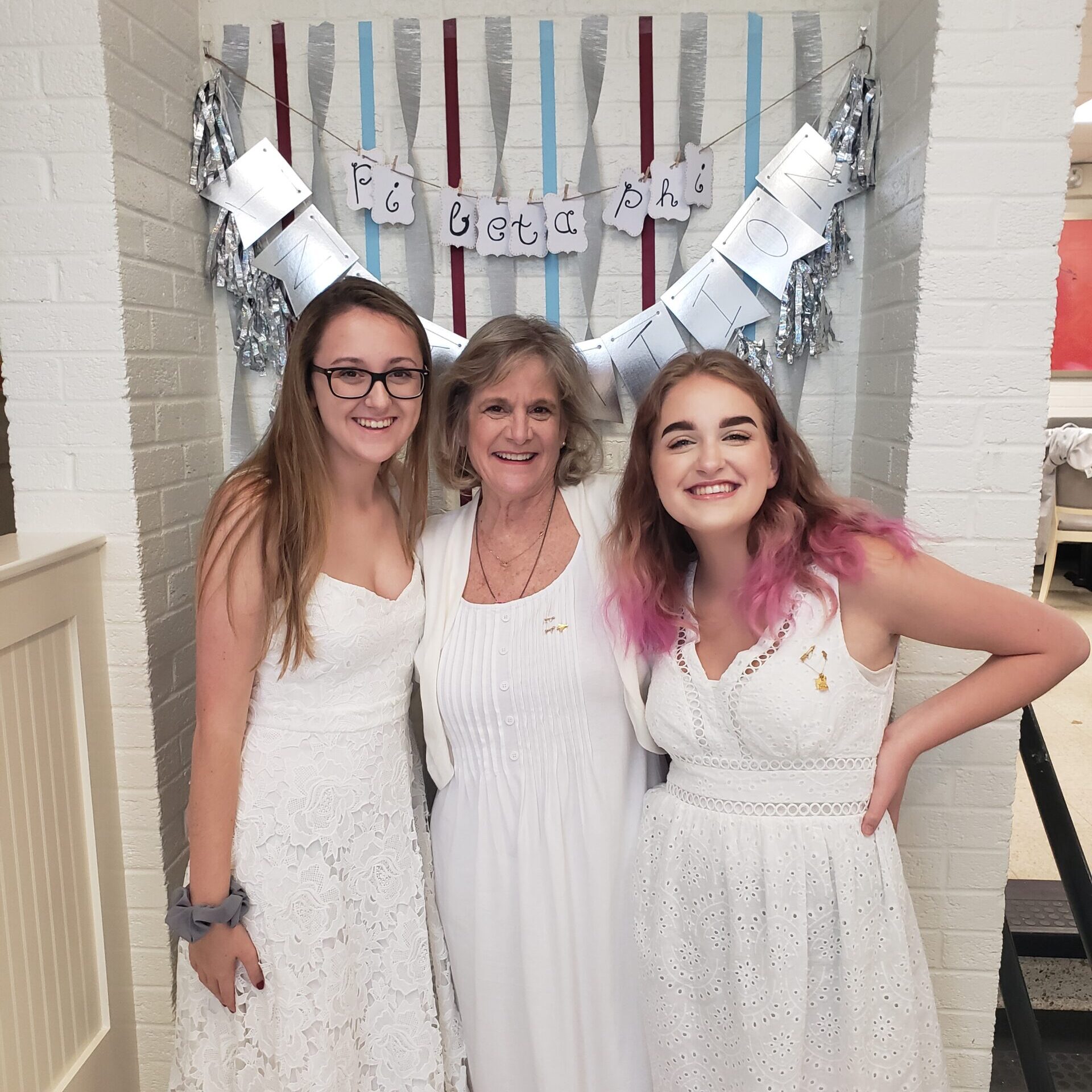 Photo of three members wearing white dresses standing in front of a wine and silver blue streamer backdrop
