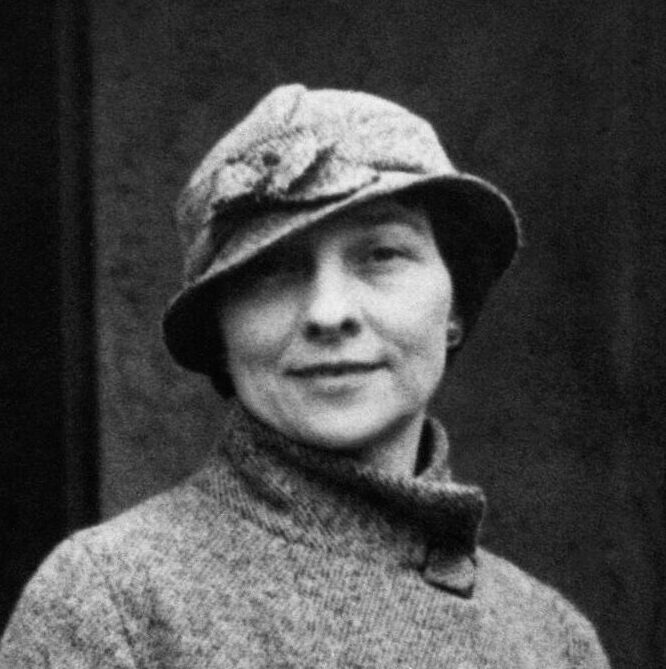 Historical black and white headshot of Elizebeth Friedman wearing a grey jacket and grey hat with a flower on it in front of a black background