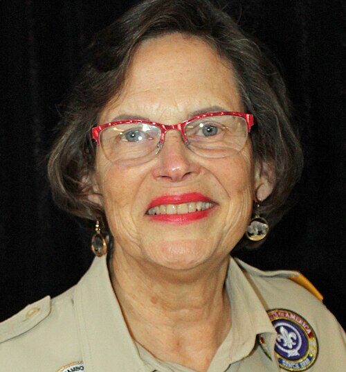 Professional headshot of Ellie Morrison wearing a Boy Scouts uniform and red glasses in front of a black background