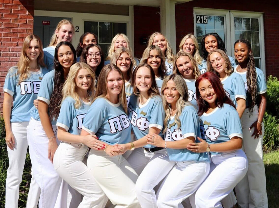 A group of individuals wearing silver blue shirts with the Pi Beta Phi Greek letters standing in front of a chapter house