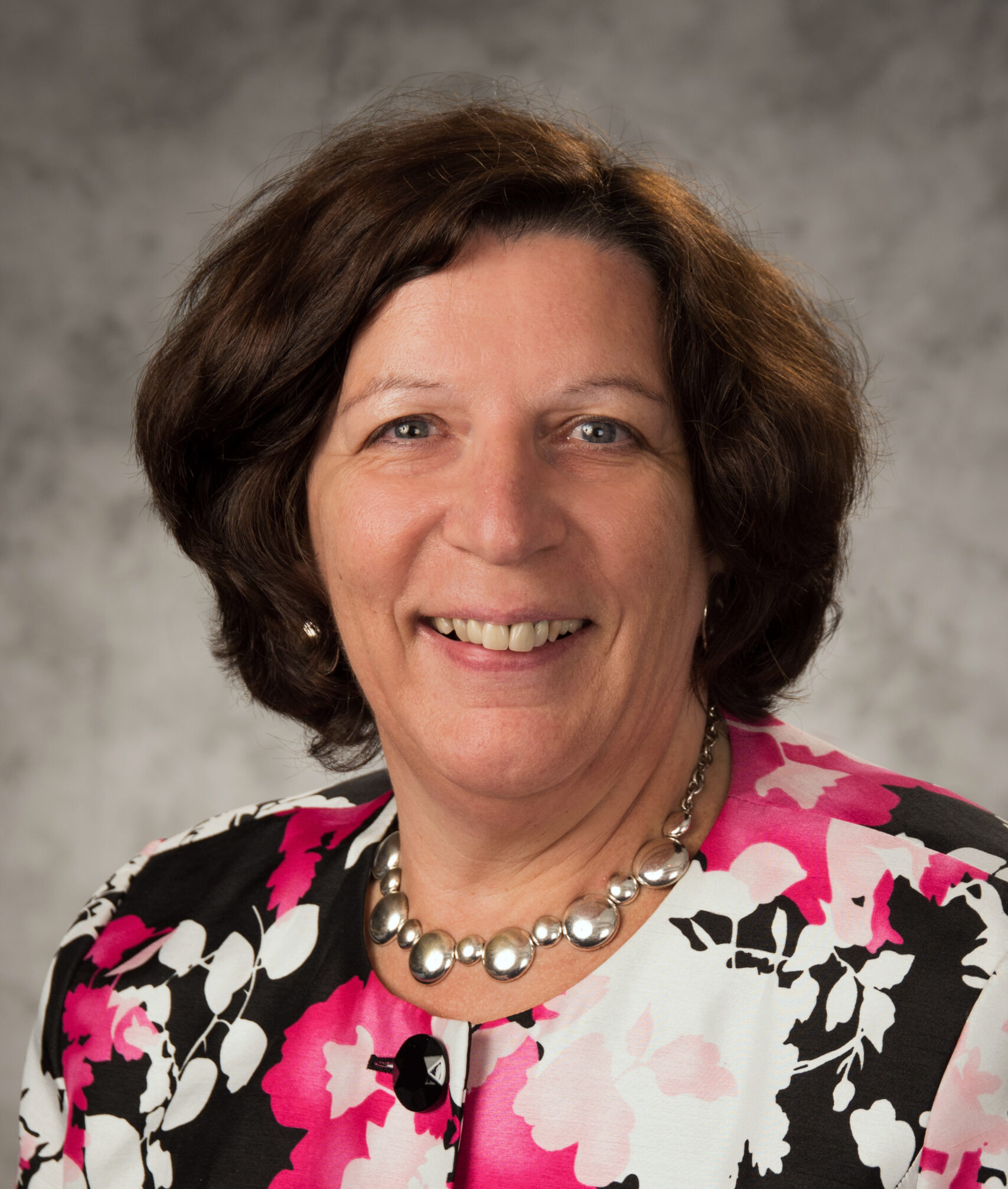 Professional headshot of Fran DeSimone Becque wearing a pink, white and black flowered shirt in front of a grey background