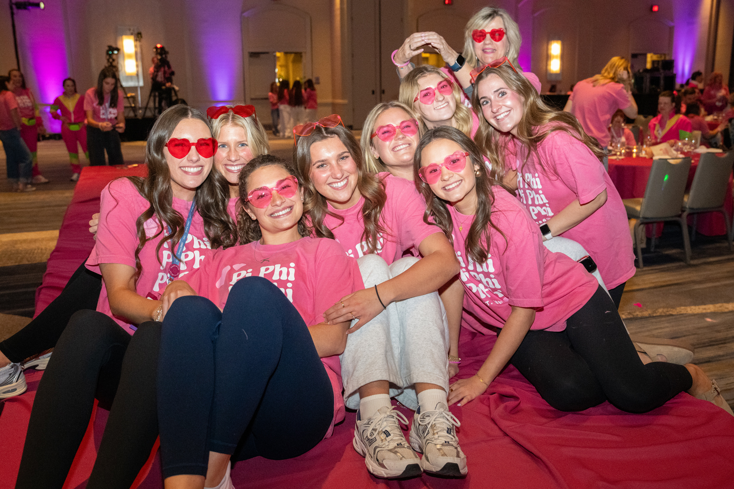 Nine members wearing pink t-shirts and heart-shaped pink sunglasses sitting together and hugging indoors on a red carpet during a conference