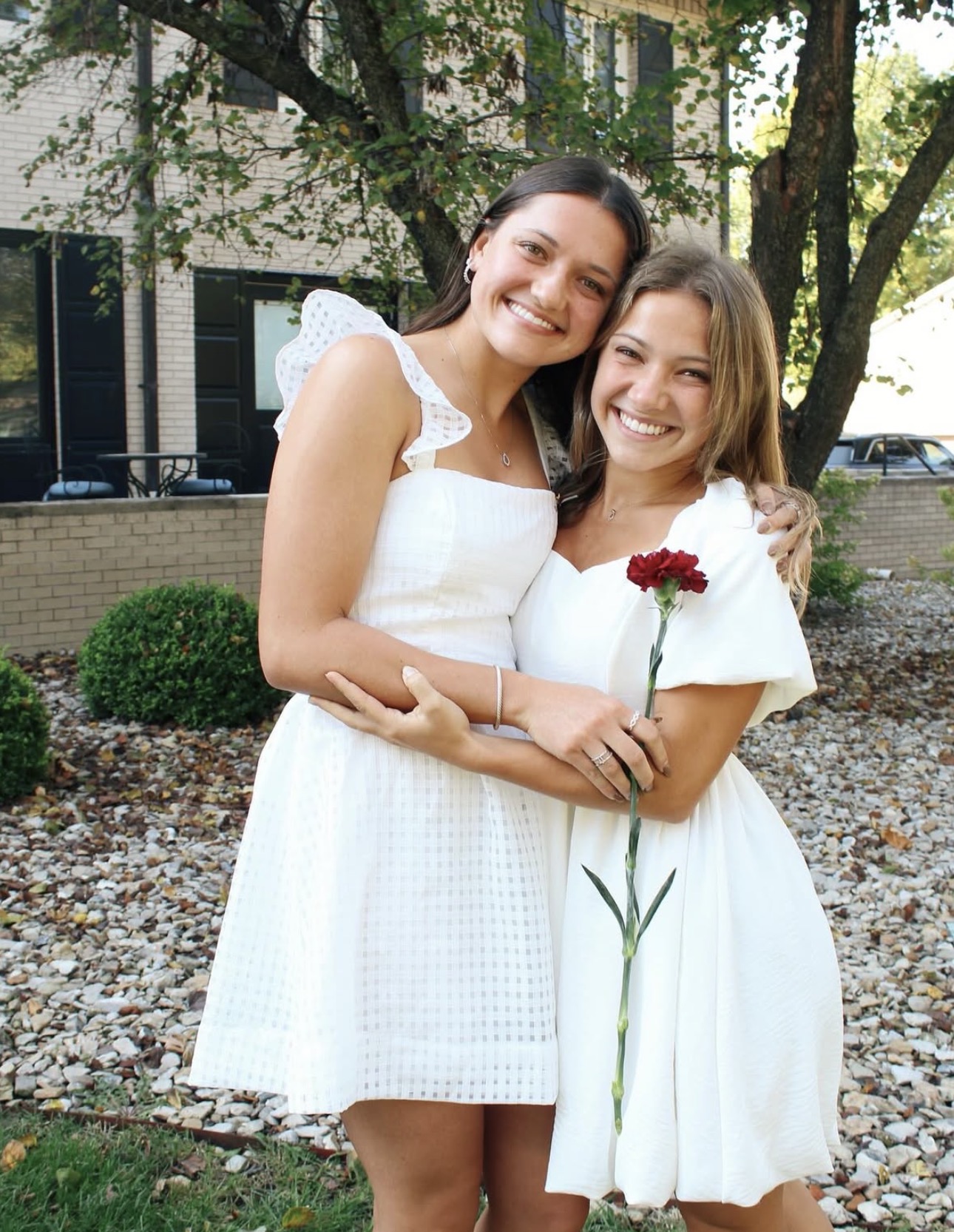 Two individuals wearing white dresses and holding a wine carnation