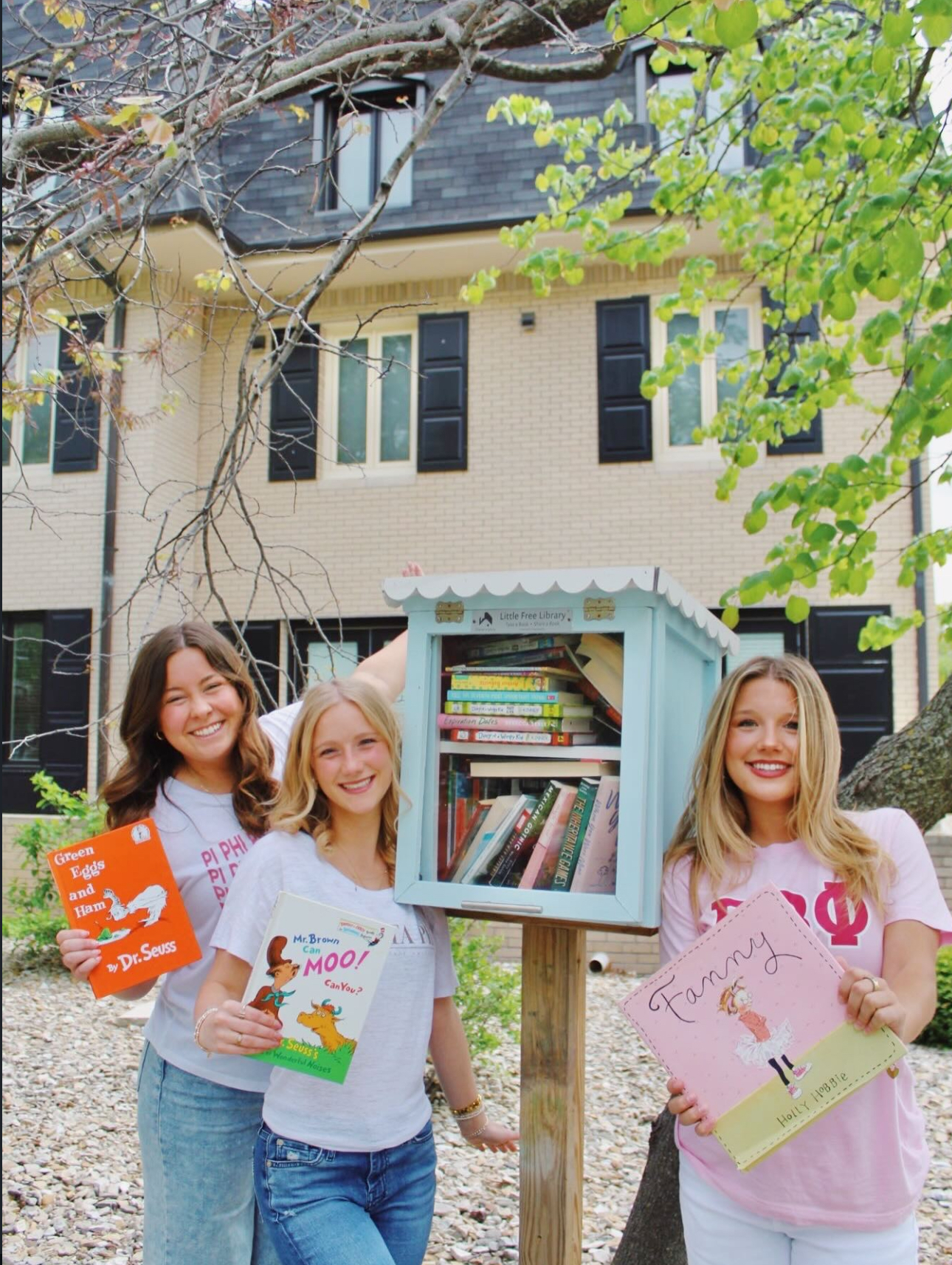 Three individuals standing besides a Little Free Library and holding children's books
