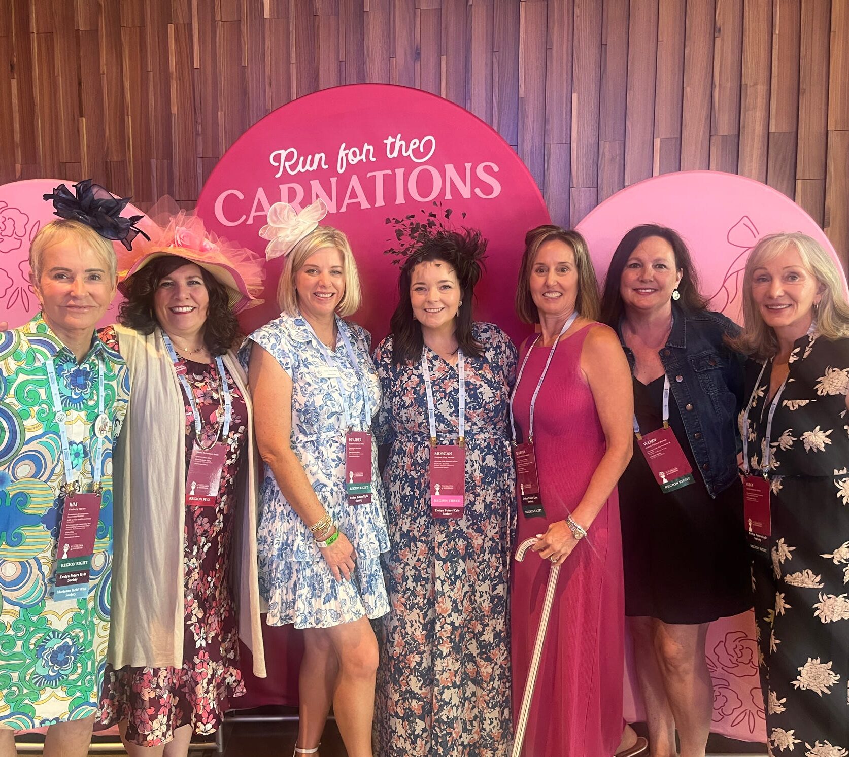 Seven members wearing fascinator hats and headbands in front of a backdrop that says Run for the Carnations