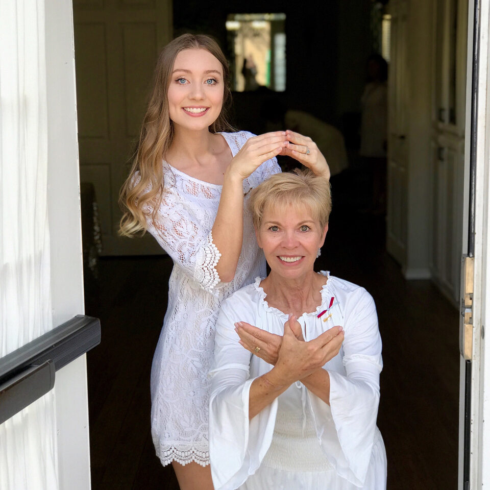 Two women wearing white; one is making a halo with their hands and holding it over the other's head. The other is making angel wings with their hands