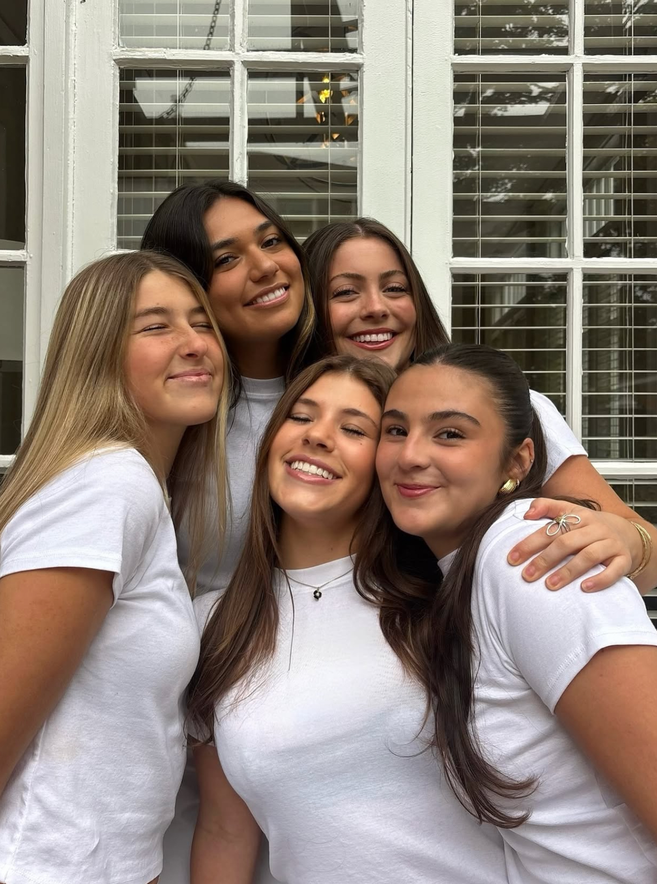 Five members wearing white t-shirts hugging outside in front of a window