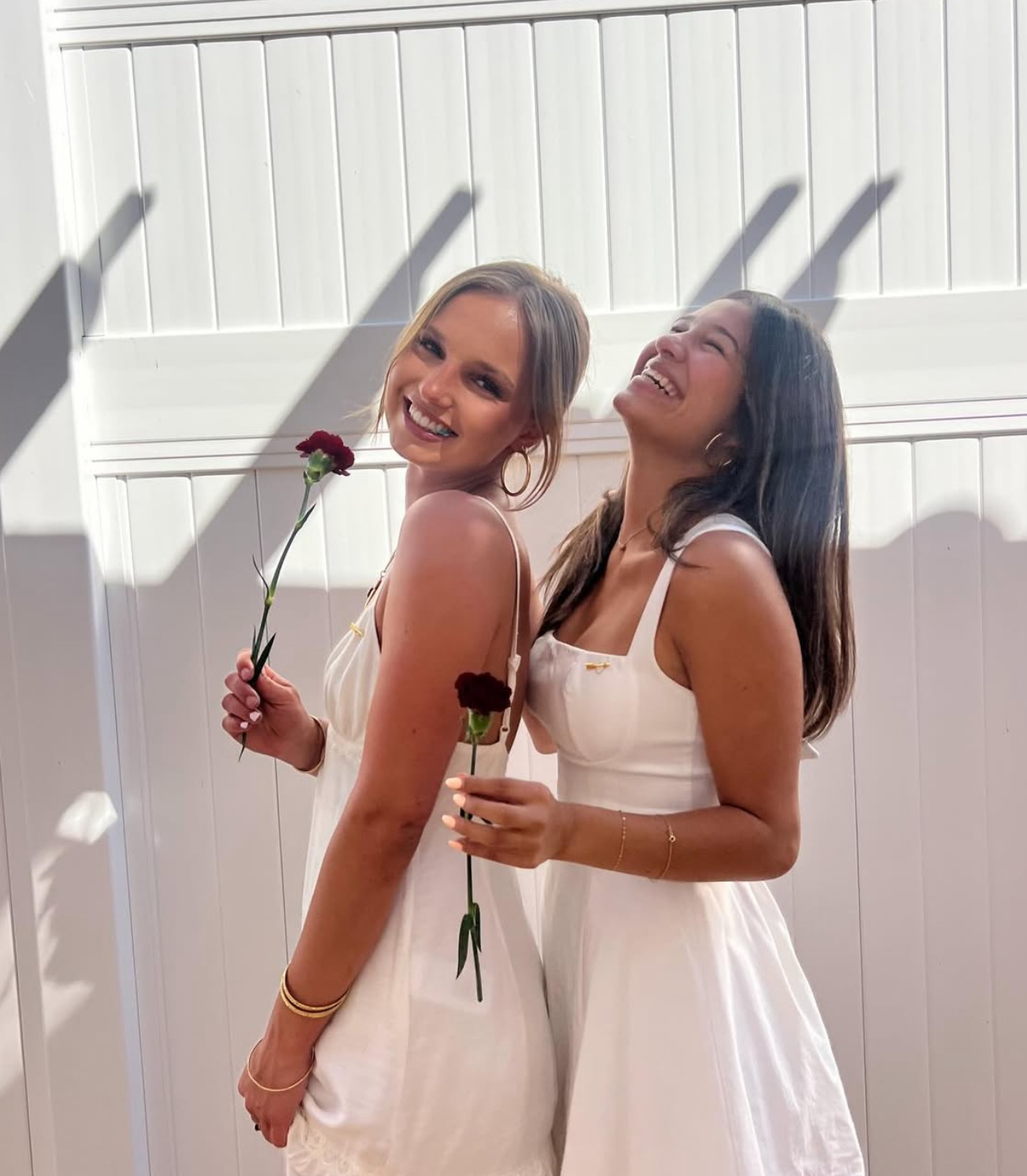 Two members wearing white dresses and holding red carnations standing outside in front of a white fence