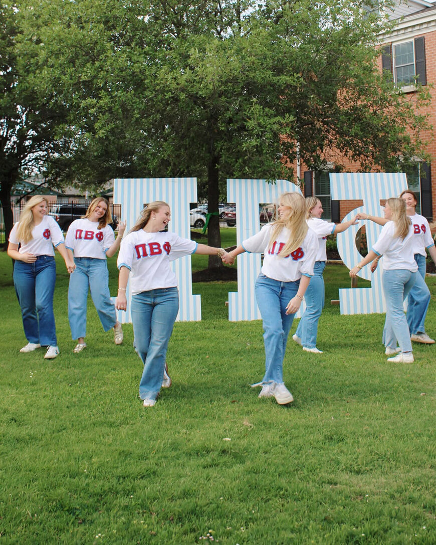 Several individuals walking in a grassy lawn in front of greek letters that say Pi Beta Phi