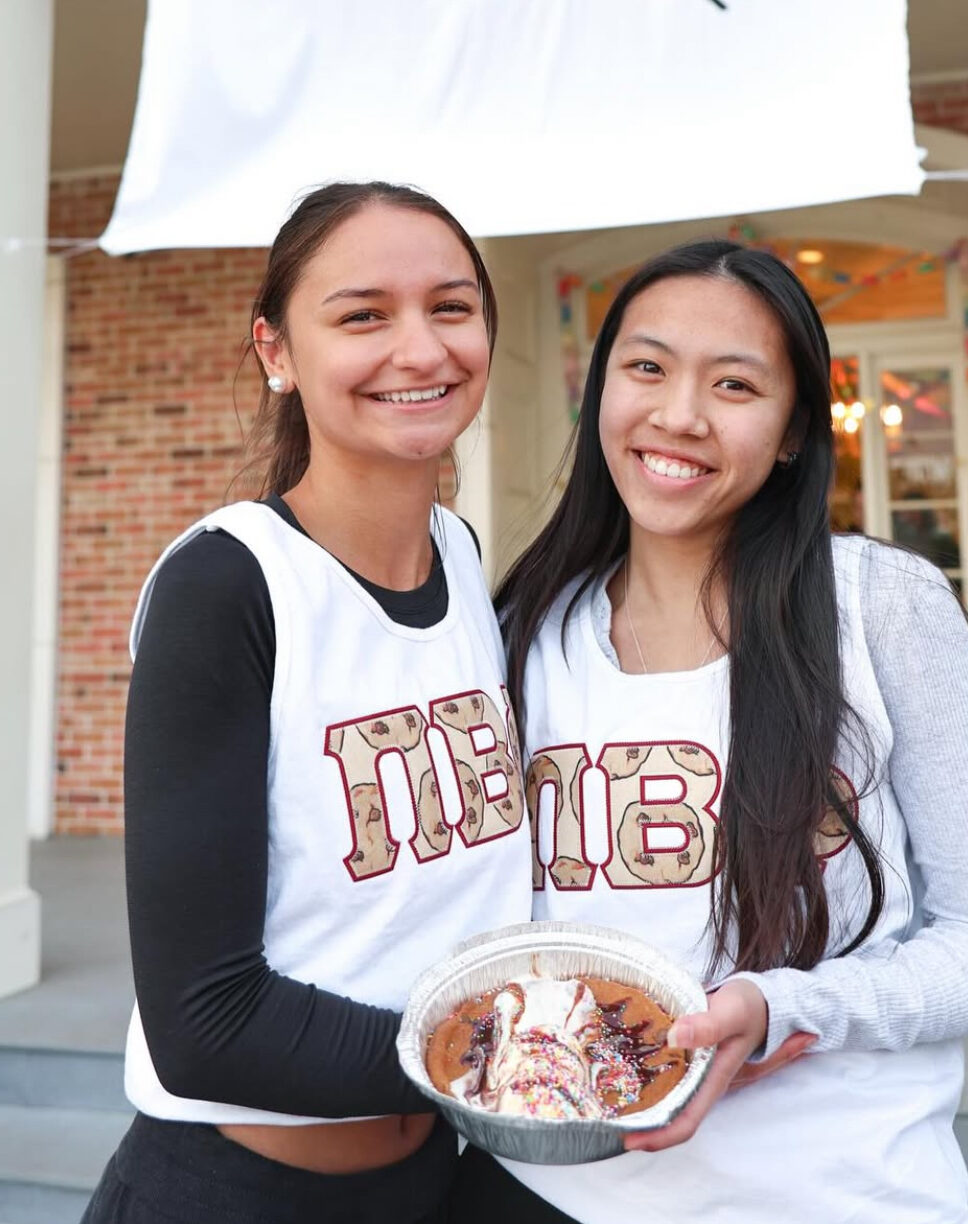 Two members wearing white jerseys with Pi Beta Phi Greek letters holding a large cookie skillet standing in front of a brick house with a white banner