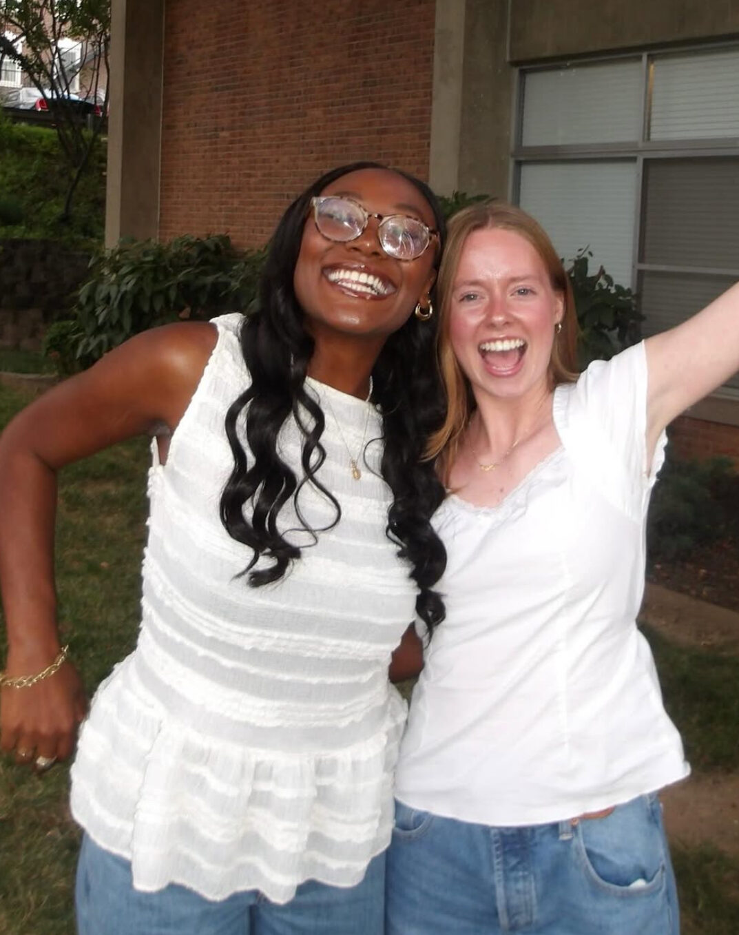Two members wearing white shirts and blue jeans hugging outside with their arms raised in front of a brick building