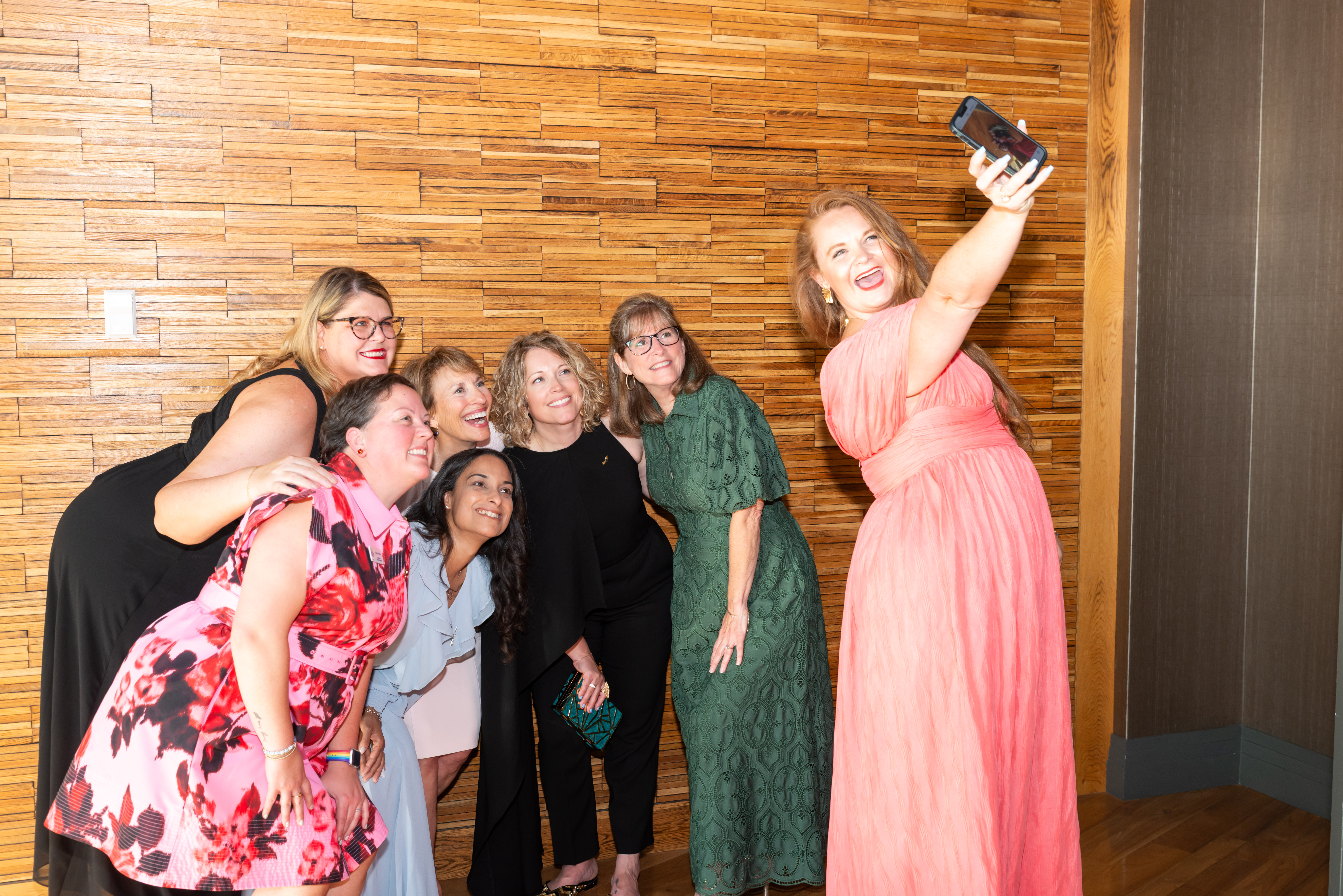 Six members pose in front of a wood wall for a selfie with one member in a pink dress holding up her phone