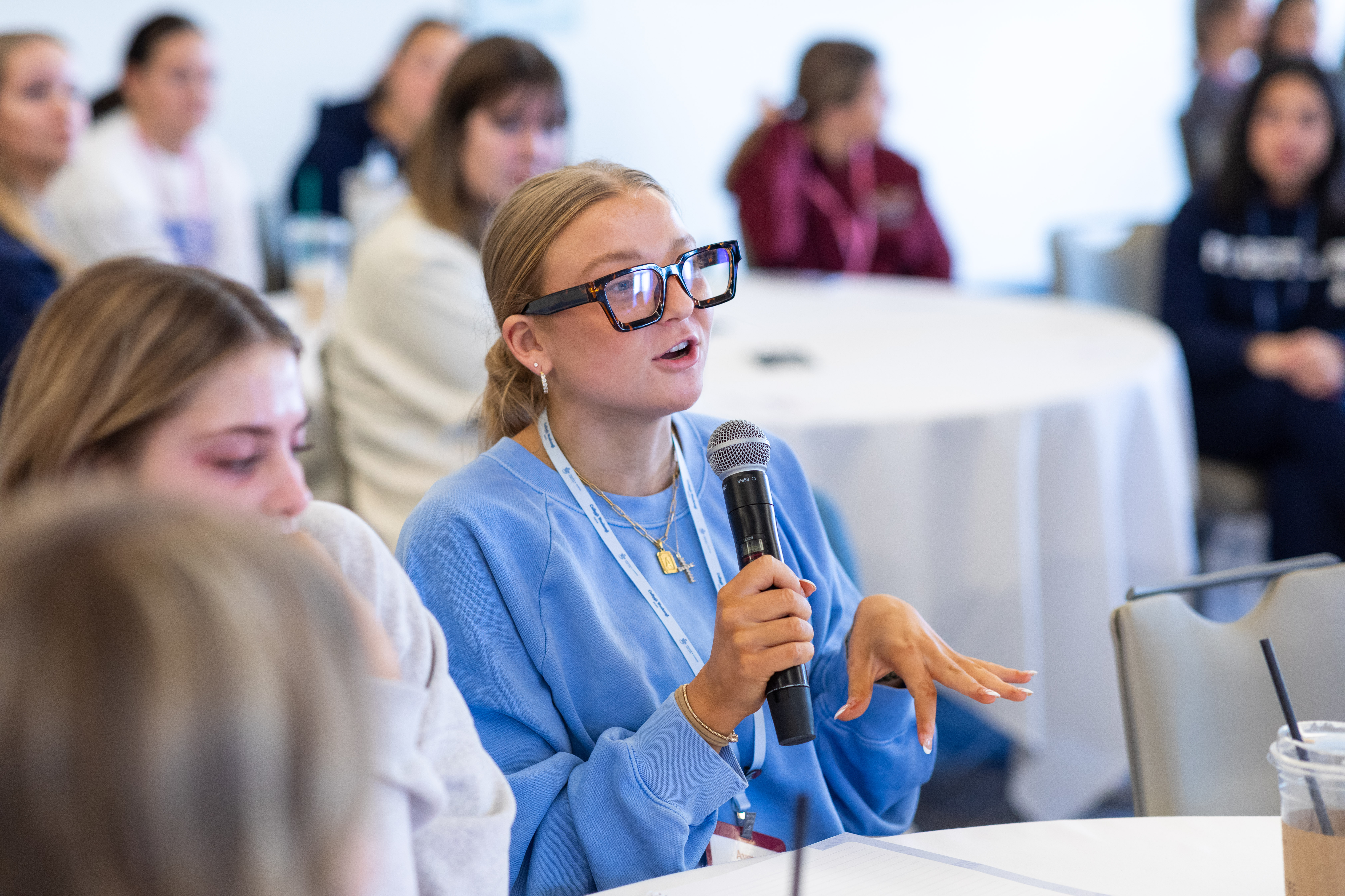 One member wearing a blue sweatshirt and glasses sitting at a table speaking into a microphone while other members listen in the background