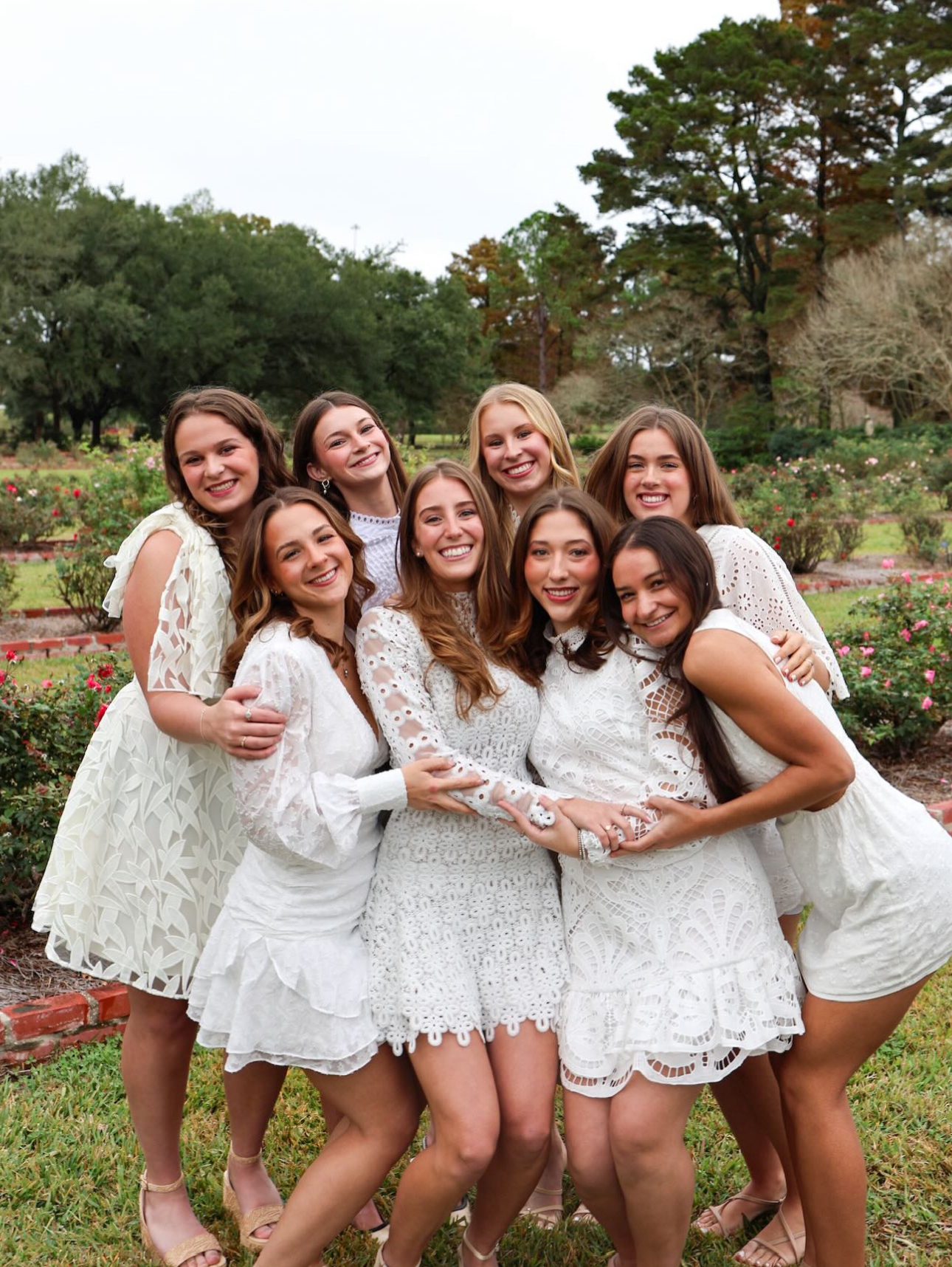 Eight members wearing white dresses hugging outside in front of green trees