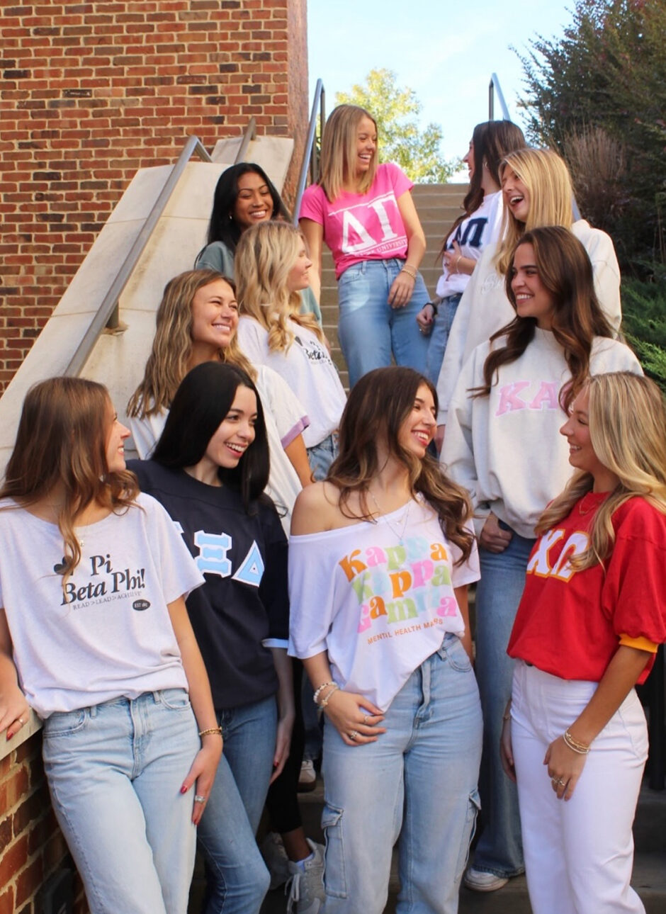Several members standing on a staircase wearing shirts with Greek letters from many organizations