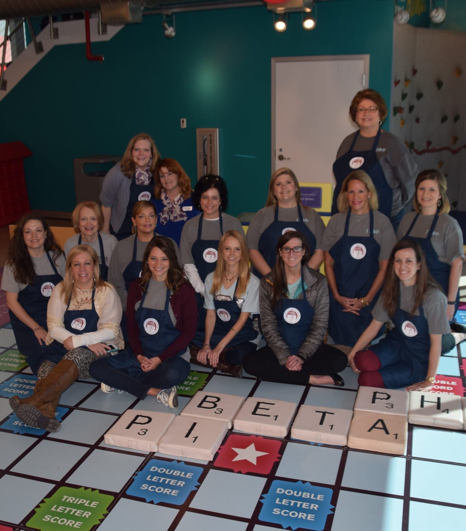 Several members wearing aprons sitting on a scrabble floor that spells Pi Beta Phi