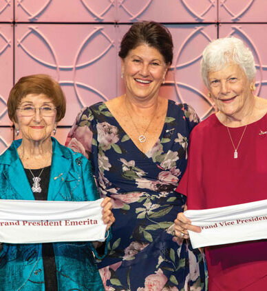 Three members standing next to one another two are holding fabric that says Grand President Emerita