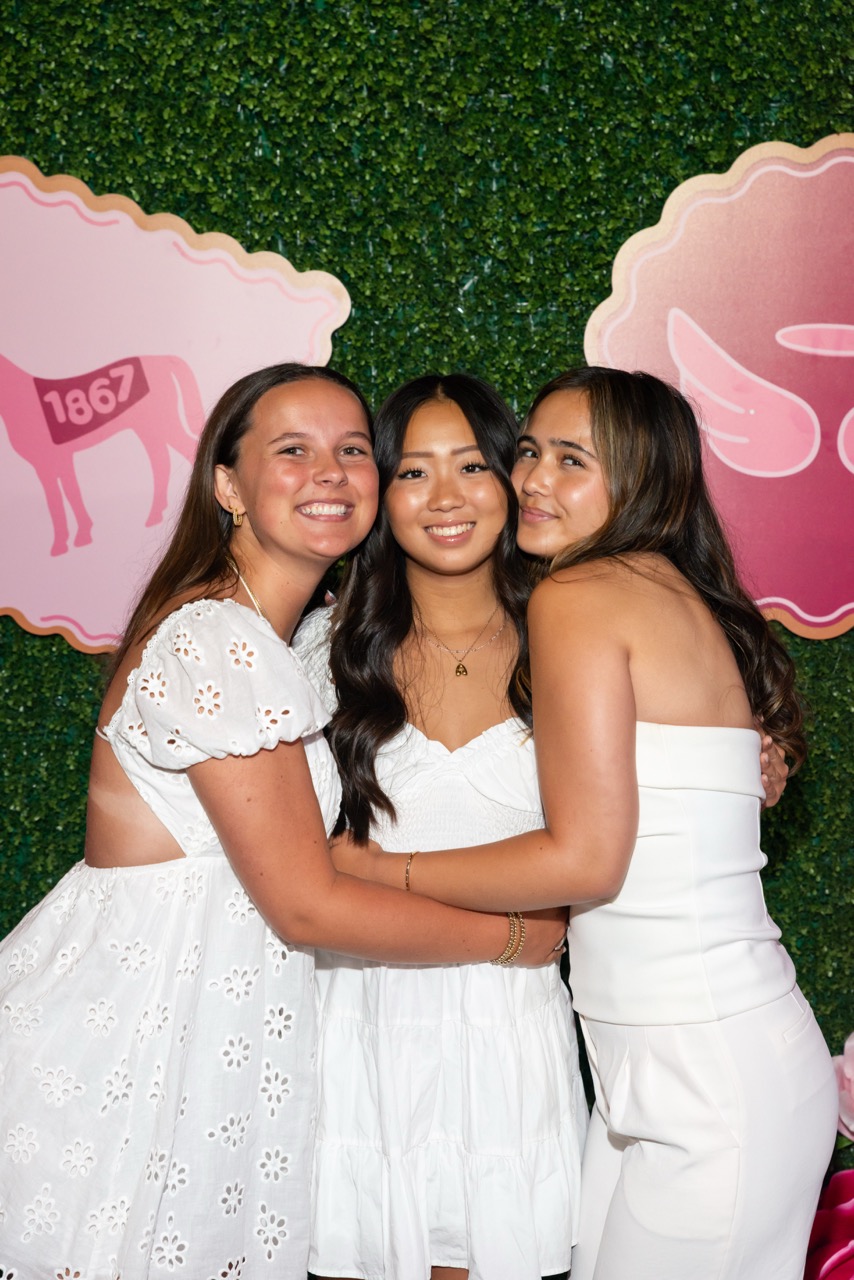 Three members wearing white standing in front of a hedge wall with pink badges