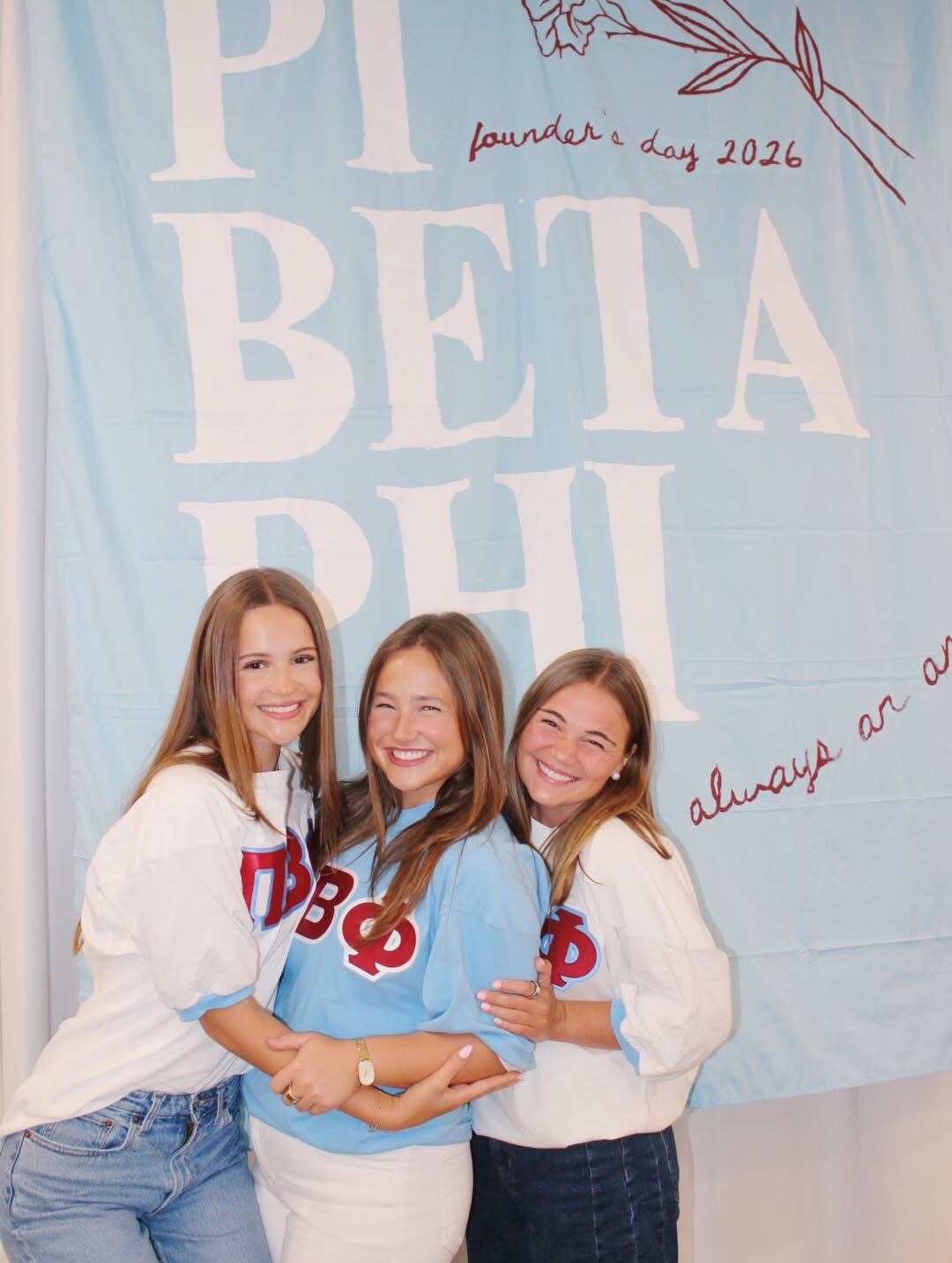 Three members hugging in front of a banner that says Pi Beta Phi Founders' Day