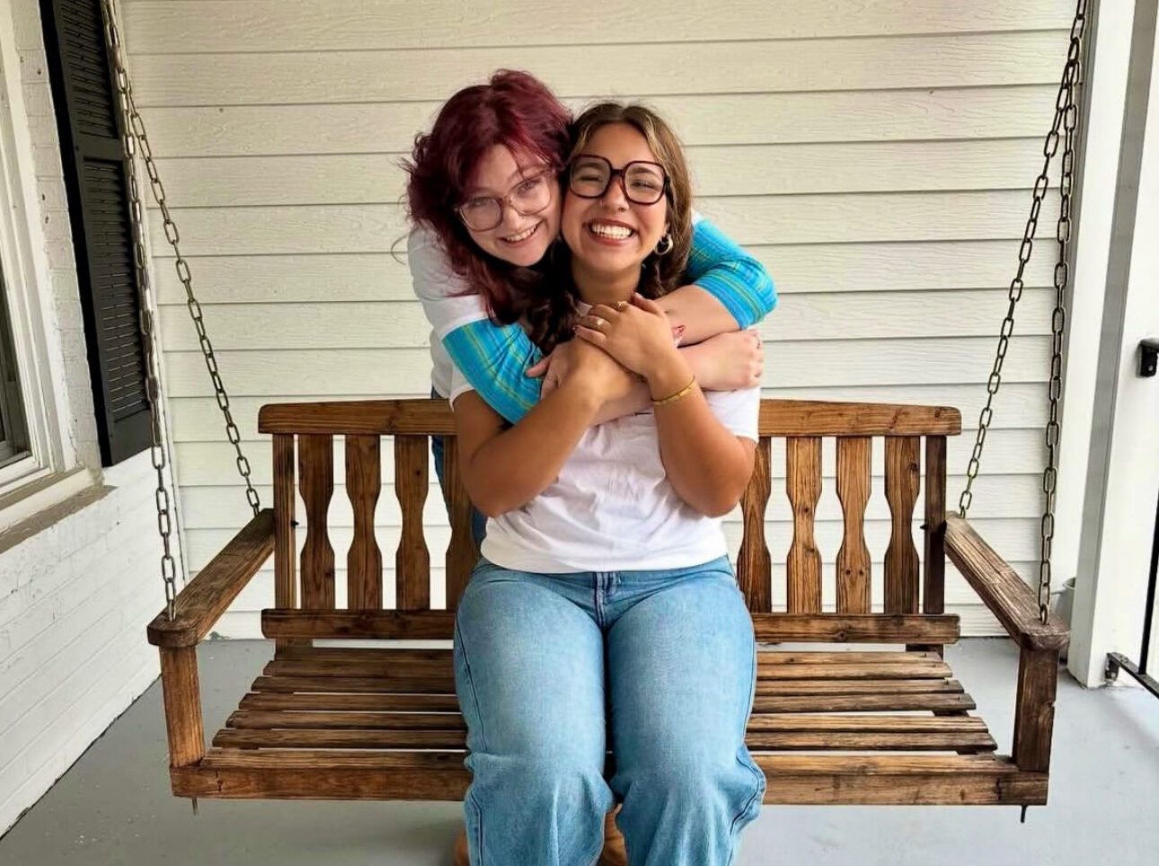 One individual sitting on a porch swing and another hugging them from behind