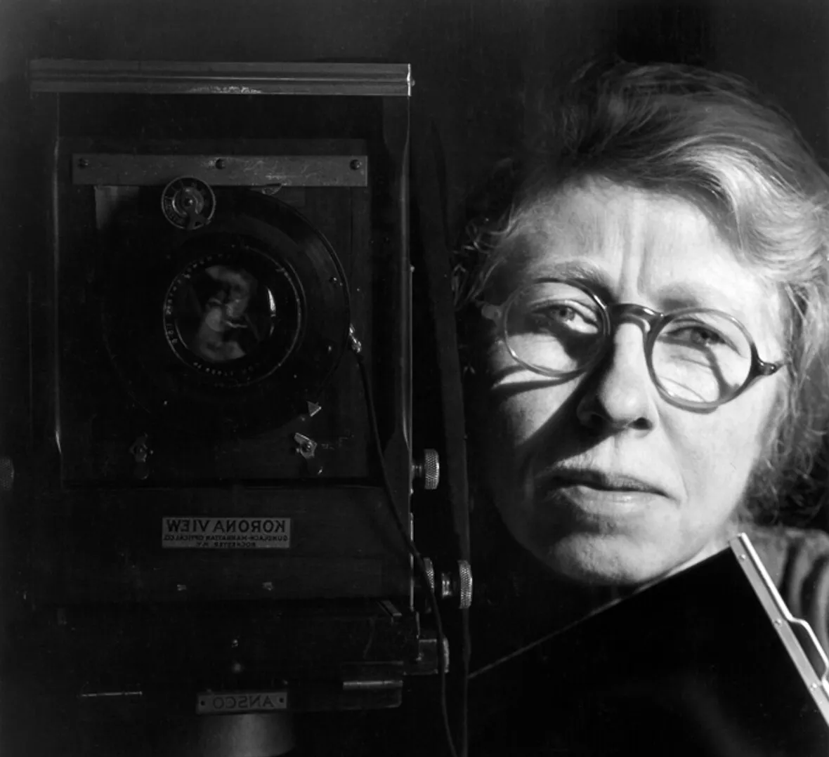 Historical black and white headshot of Imogen Cunningham wearing a black shirt and glasses in front of a black backgrounf