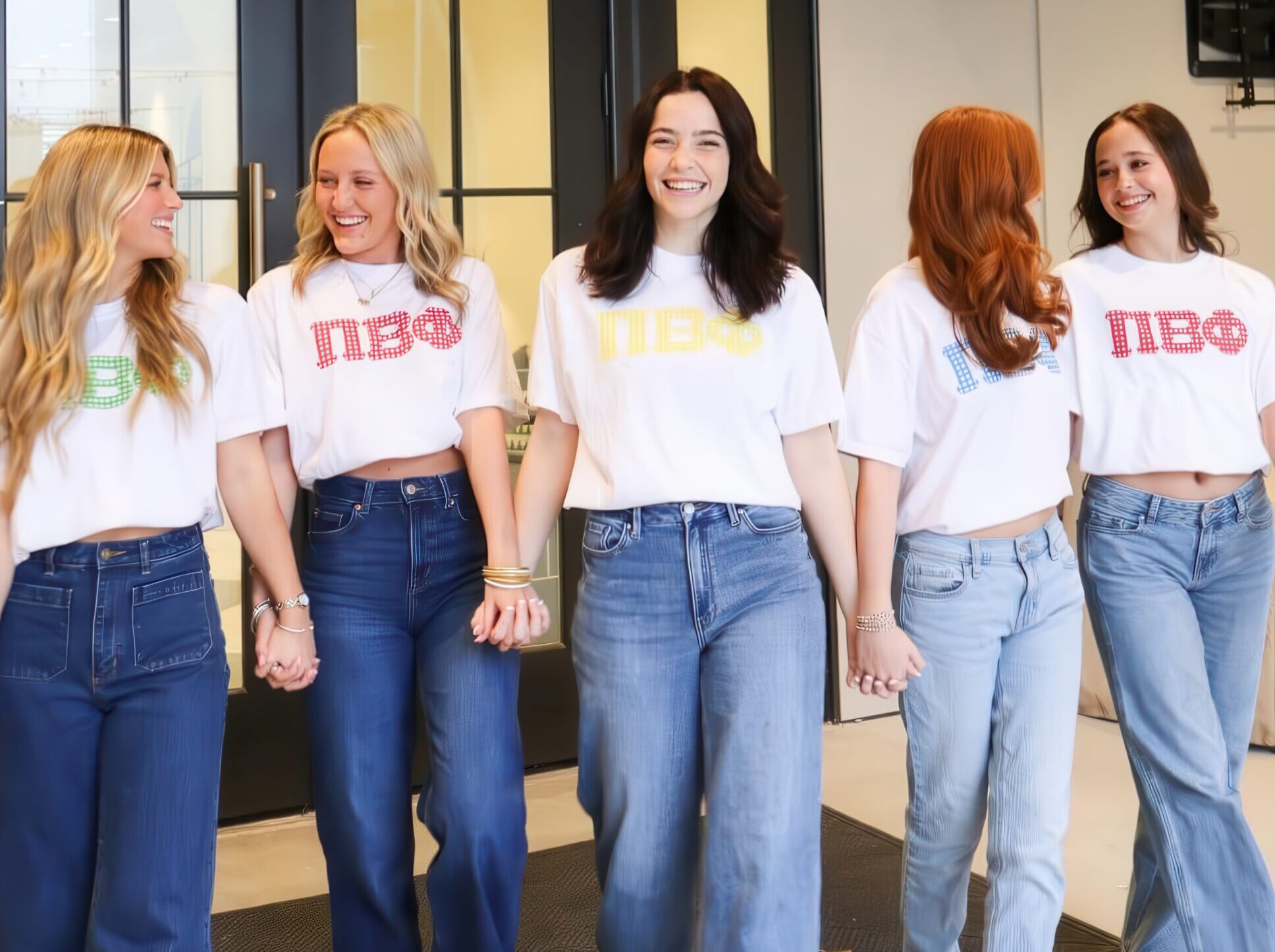 Five Pi Beta Phi members laughing together in front of a chapter house entrance, wearing white Pi Beta Phi Greek letter t-shirts in different colors with jeans